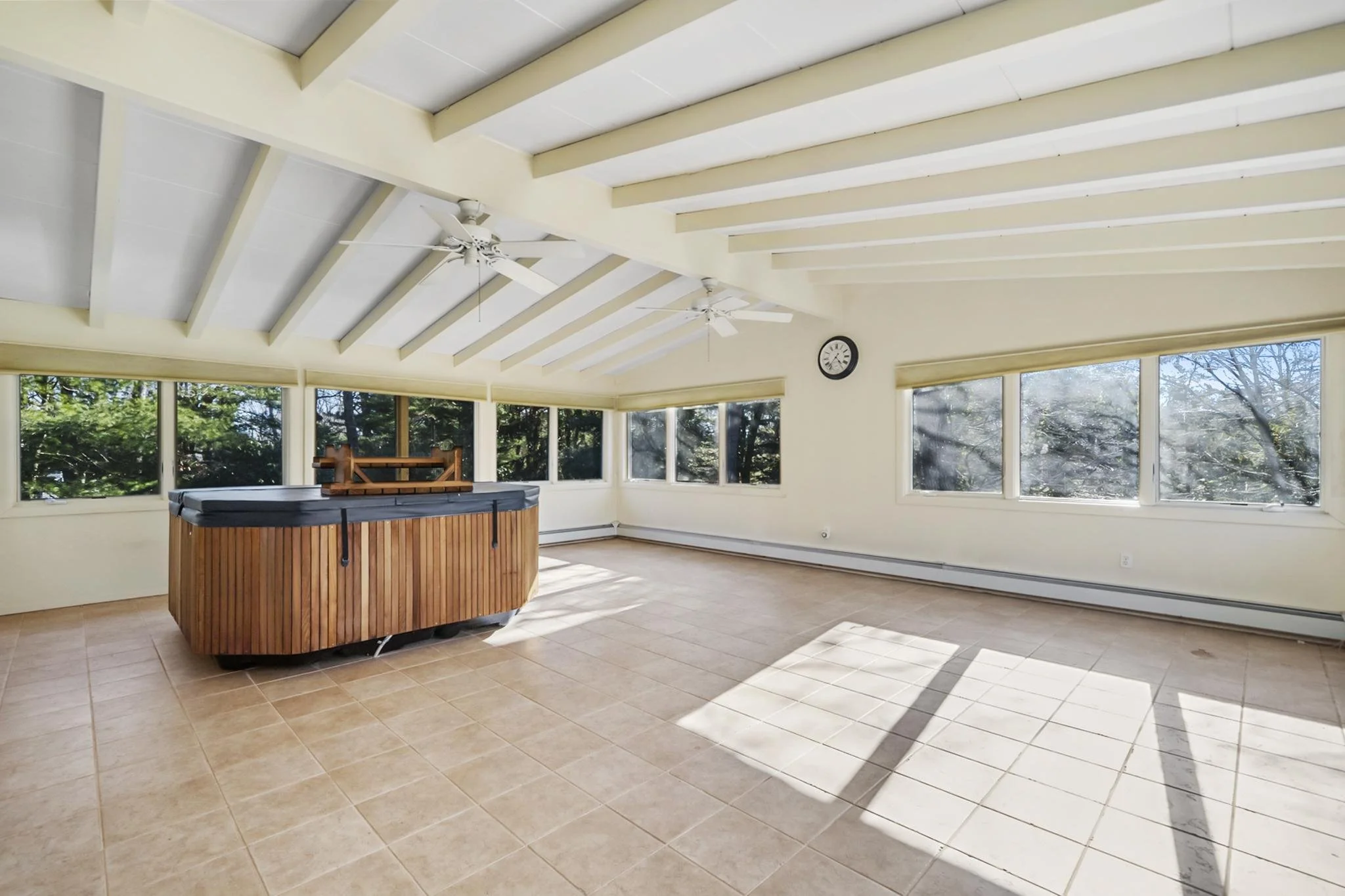 An indoor sunroom with large windows, tiled floor, ceiling fans, a round hot tub, and a clock on the wall, with trees visible outside.