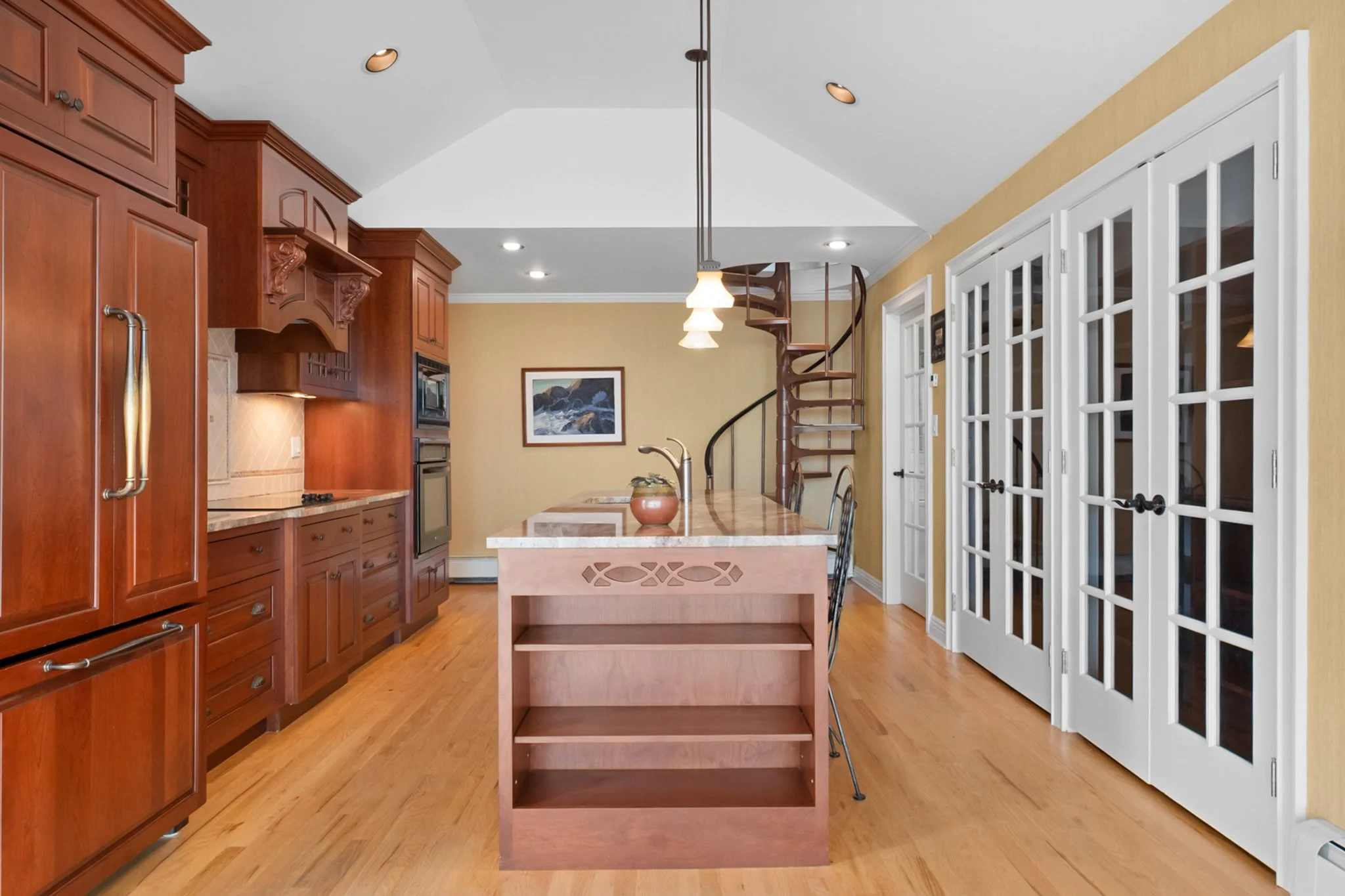 Kitchen with wooden cabinets, a central island, white French doors on the right, a spiral staircase in the back, and warm lighting.