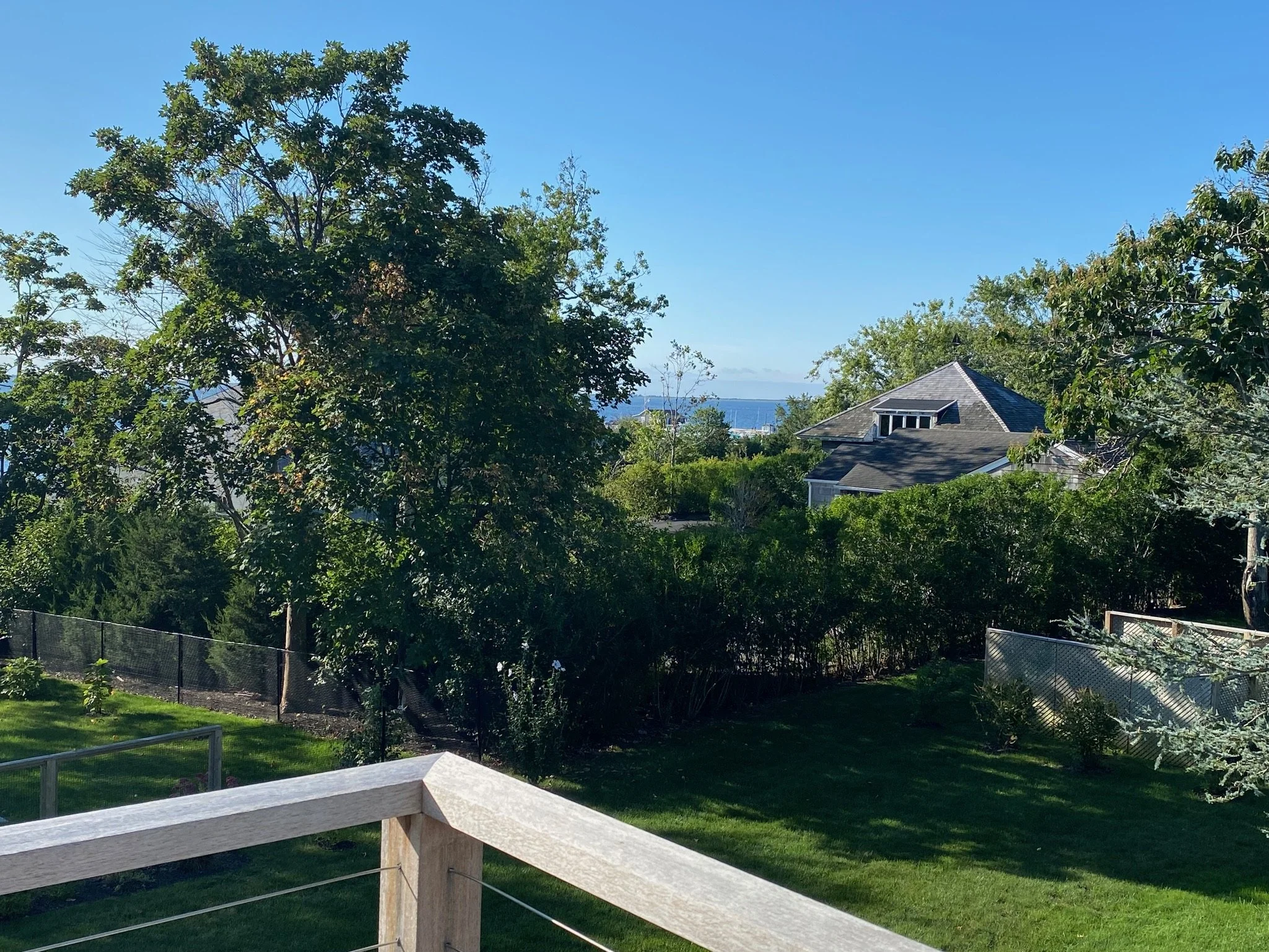 A backyard with a wooden deck railing, a fenced garden area with plants, trees, a house with a dark roof, and beyond that, trees and the ocean under a clear blue sky.