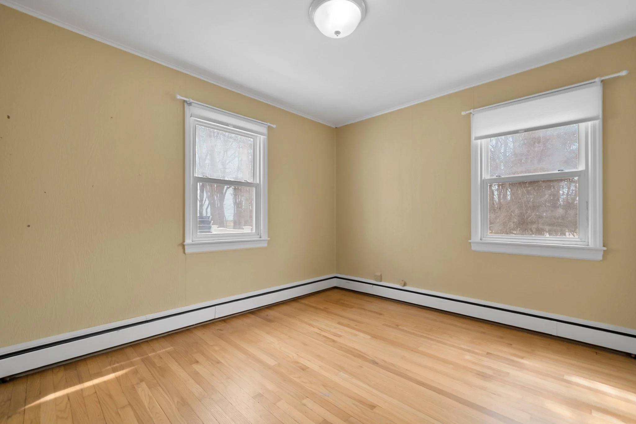 Empty room with light yellow walls, two windows with white blinds, hardwood floor, and white ceiling with a ceiling light fixture.