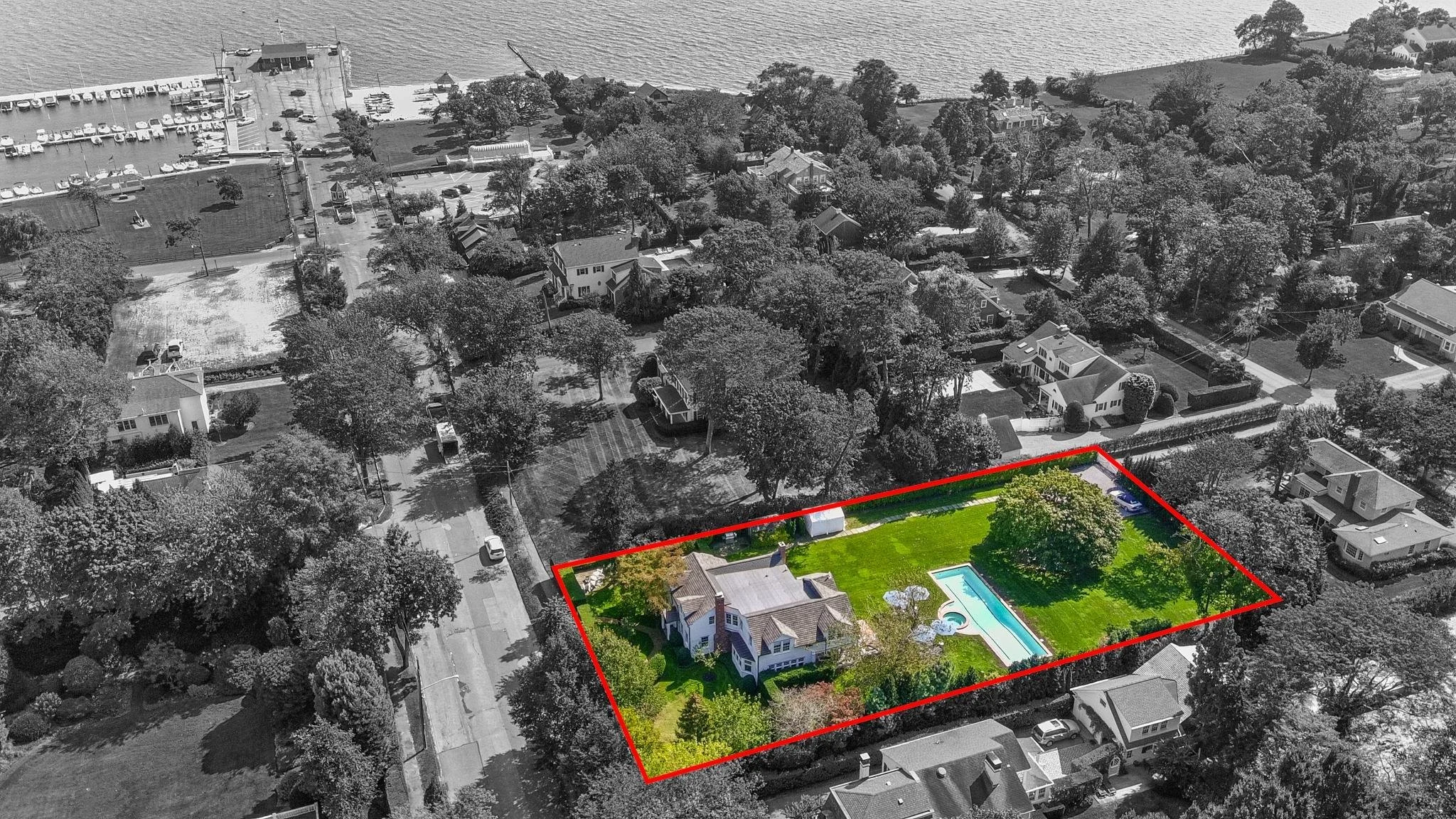 Aerial view of a neighborhood with trees and houses. A red outline highlights a property with a house, a swimming pool, and a lawn.