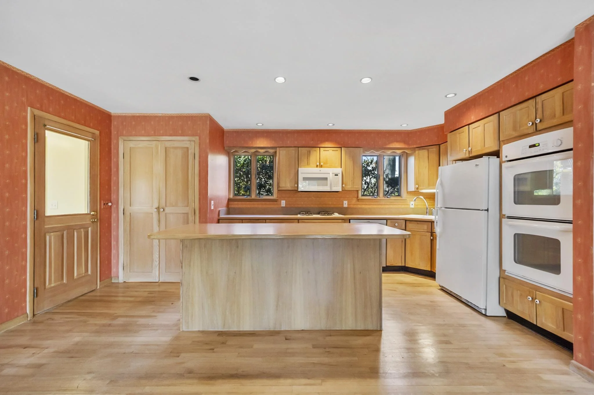 Kitchen with wooden cabinets, white appliances, a kitchen island, and two small windows showing trees outside.