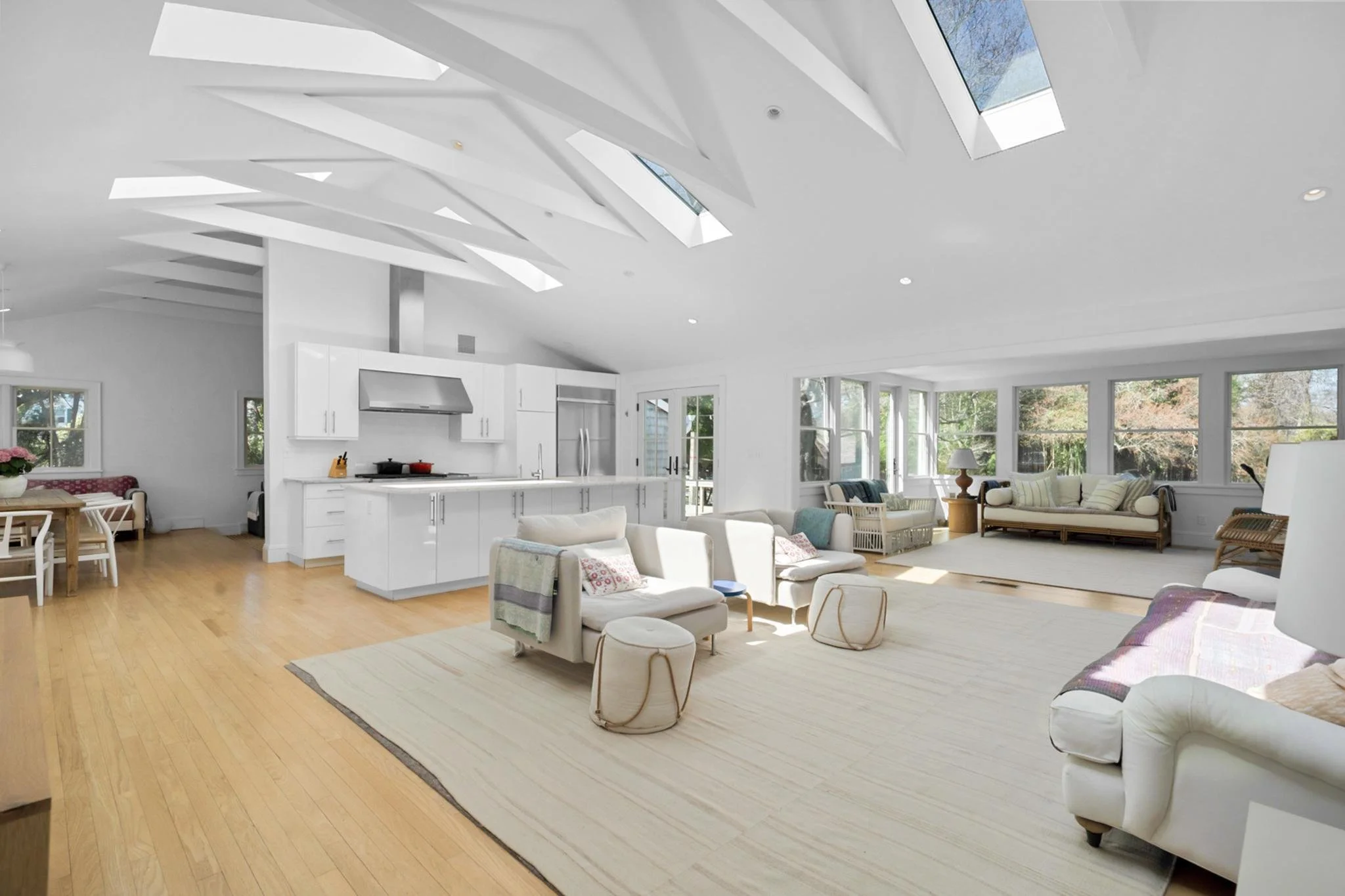 Spacious living room with white walls, large windows, and vaulted ceiling featuring skylights. Furnished with white sofas, armchairs, a wooden dining table with chairs, a kitchen with white cabinets, and hardwood floors. Natural light fills the room,