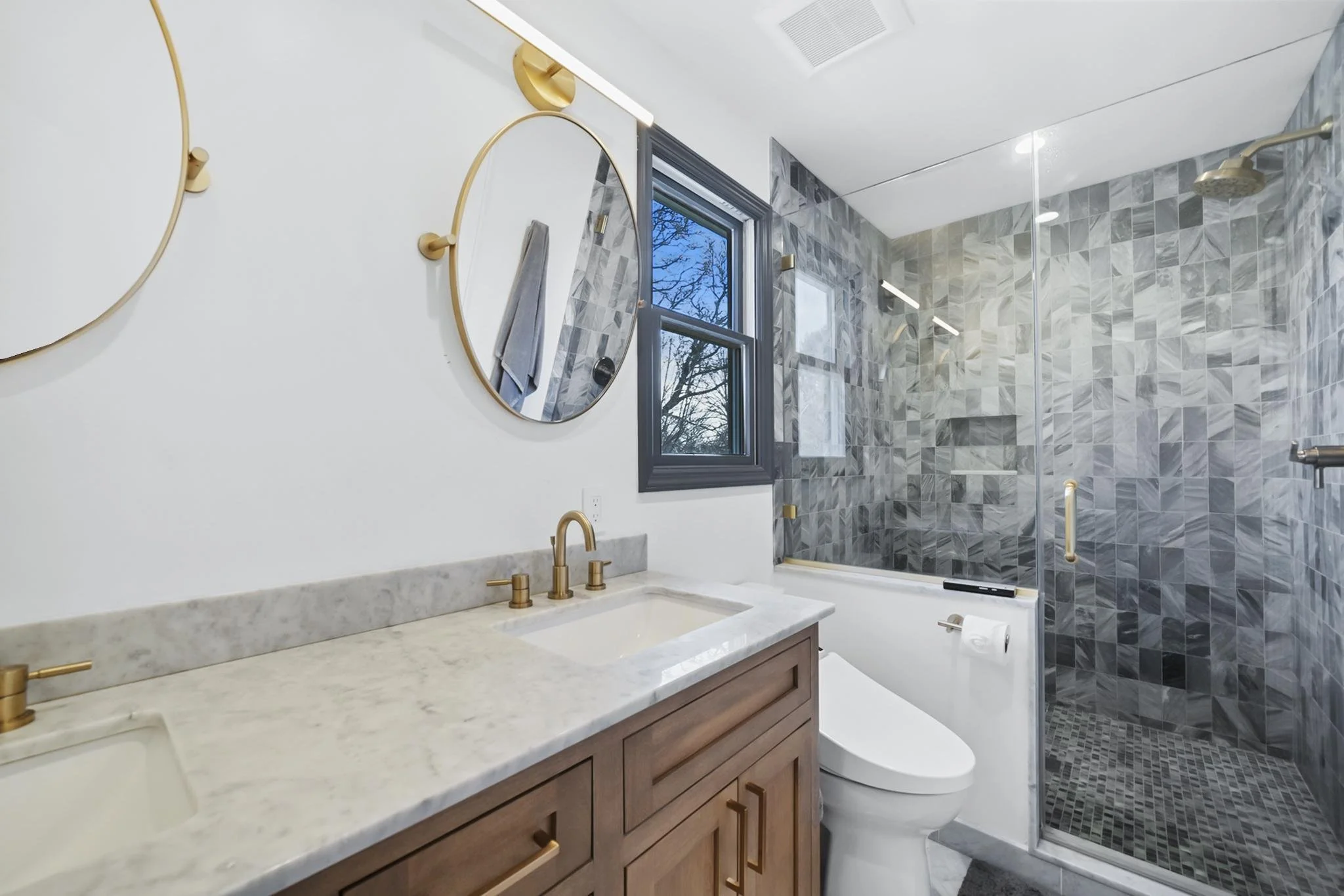 Modern bathroom with a wooden vanity, marble countertop, and gold fixtures. There are two circular mirrors on the wall, a window showing trees outside, and a marble-tiled shower with a glass door and gold accents.
