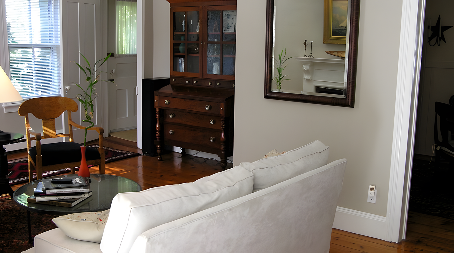 Living room with beige sofa, glass coffee table with books, wooden chair, and a tall plant in an orange vase. There is a mirror on the wall and a wooden display cabinet with glass doors. The room has wooden flooring and natural light from windows.