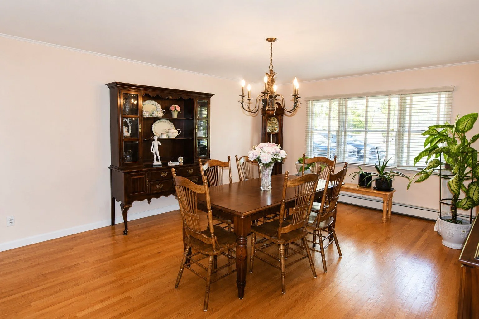 Dining room with wooden table, six chairs, a floral centerpiece, a dark wood china cabinet, an antique clock, potted plants, and a large window with wooden blinds.