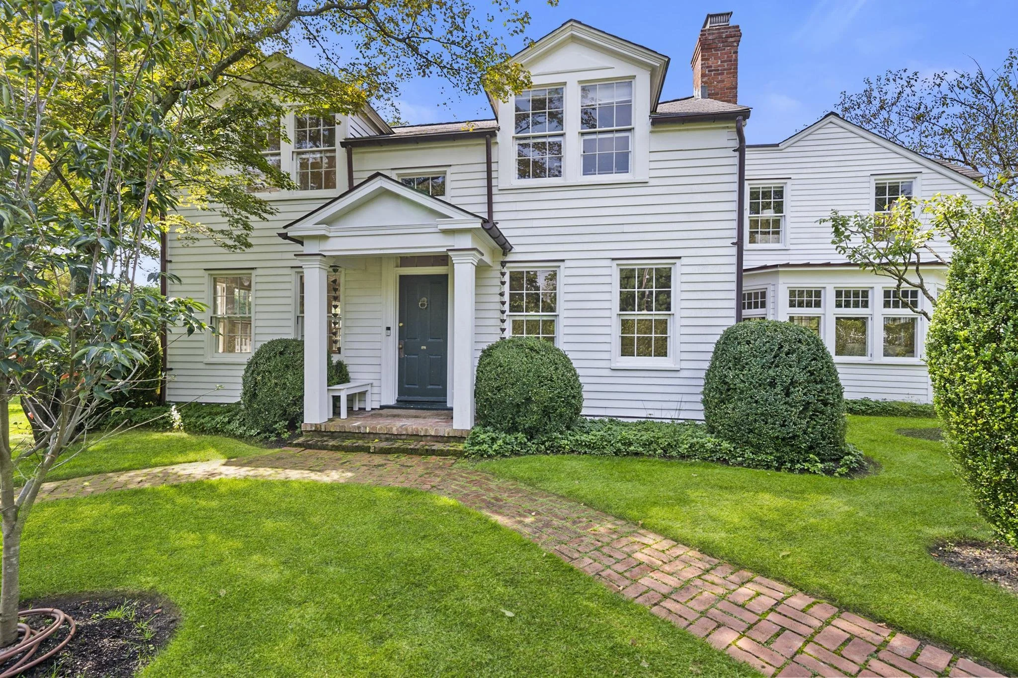 White two-story house with multiple windows, a blue front door, brick steps, and surrounding green lawn and bushes under blue sky.