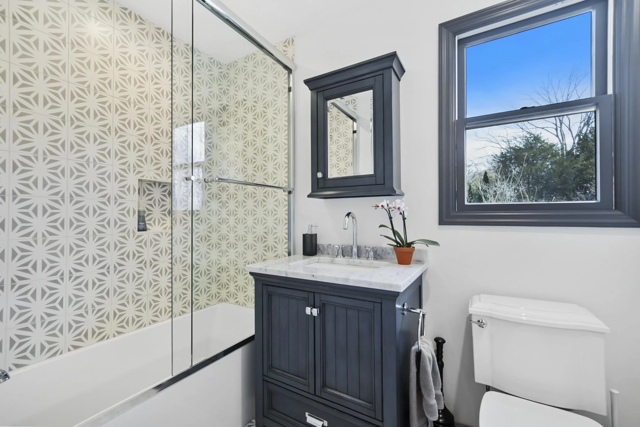 A modern bathroom with a shower area featuring patterned beige tiles, a dark blue vanity with a marble countertop, a small mirror cabinet, a white toilet, a black toilet brush holder, and two large windows showing a clear blue sky and trees outside.