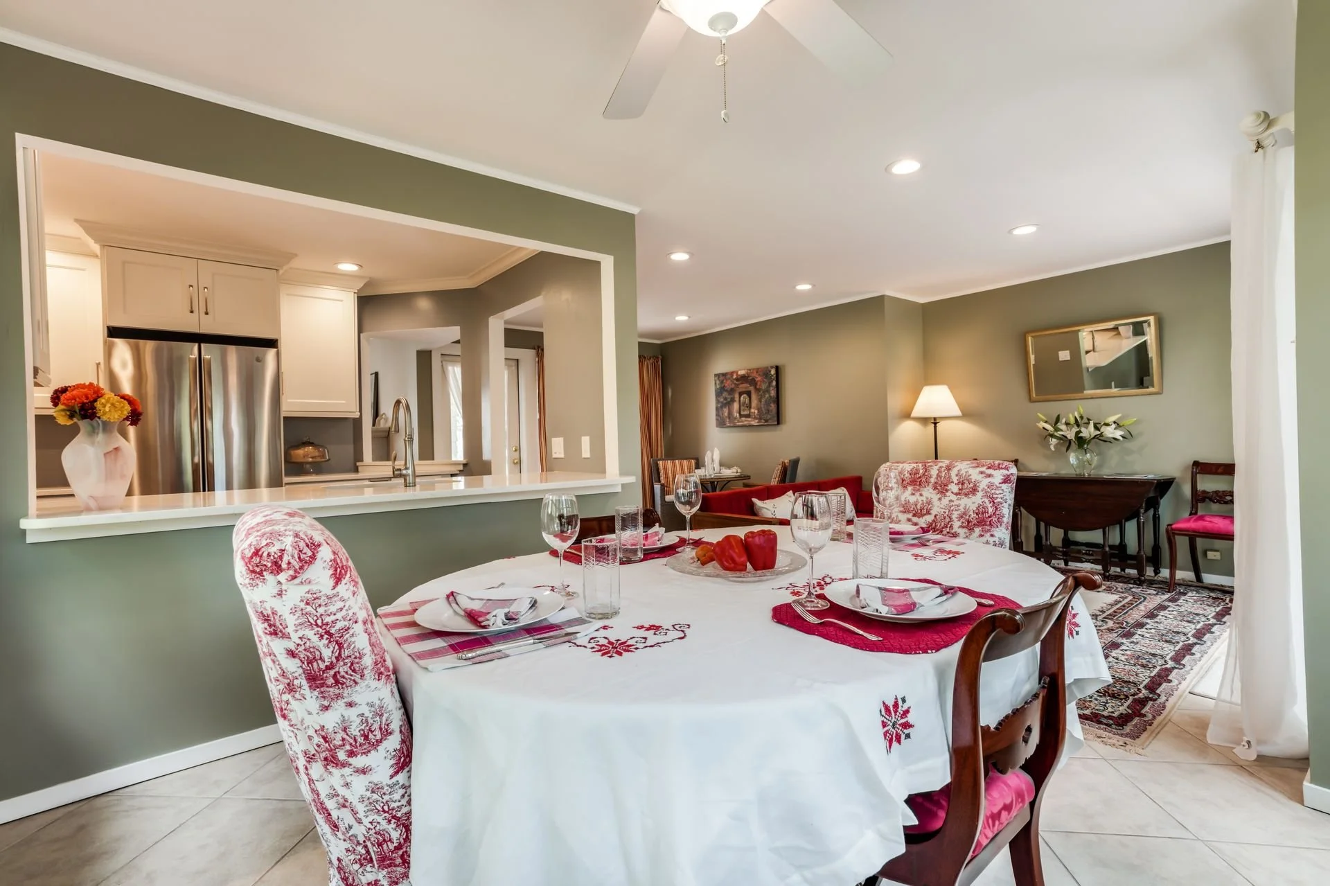 A dining room with a round table set for a meal, with red and white chairs, red and white tablecloth, and glassware and table decor. The background shows a living space with green walls, a lamp, painting, mirror, plants, and furniture.