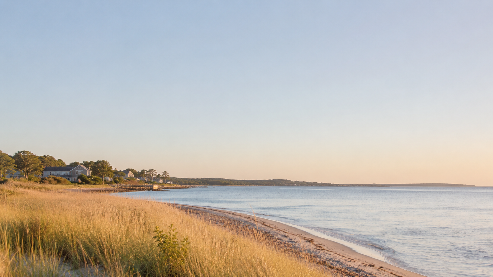 A peaceful beach scene with golden sand, calm water, houses on the shoreline, grassy dunes, and a clear sky during sunset or sunrise.