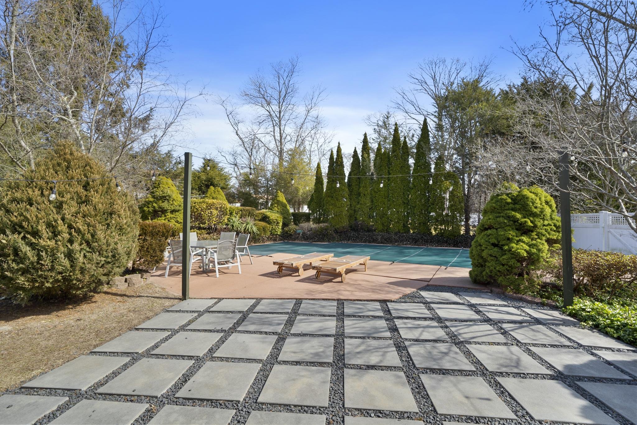 A backyard patio scene with outdoor furniture, a swimming pool with a cover, surrounded by trees and bushes, with string lights hanging above, under a clear blue sky.