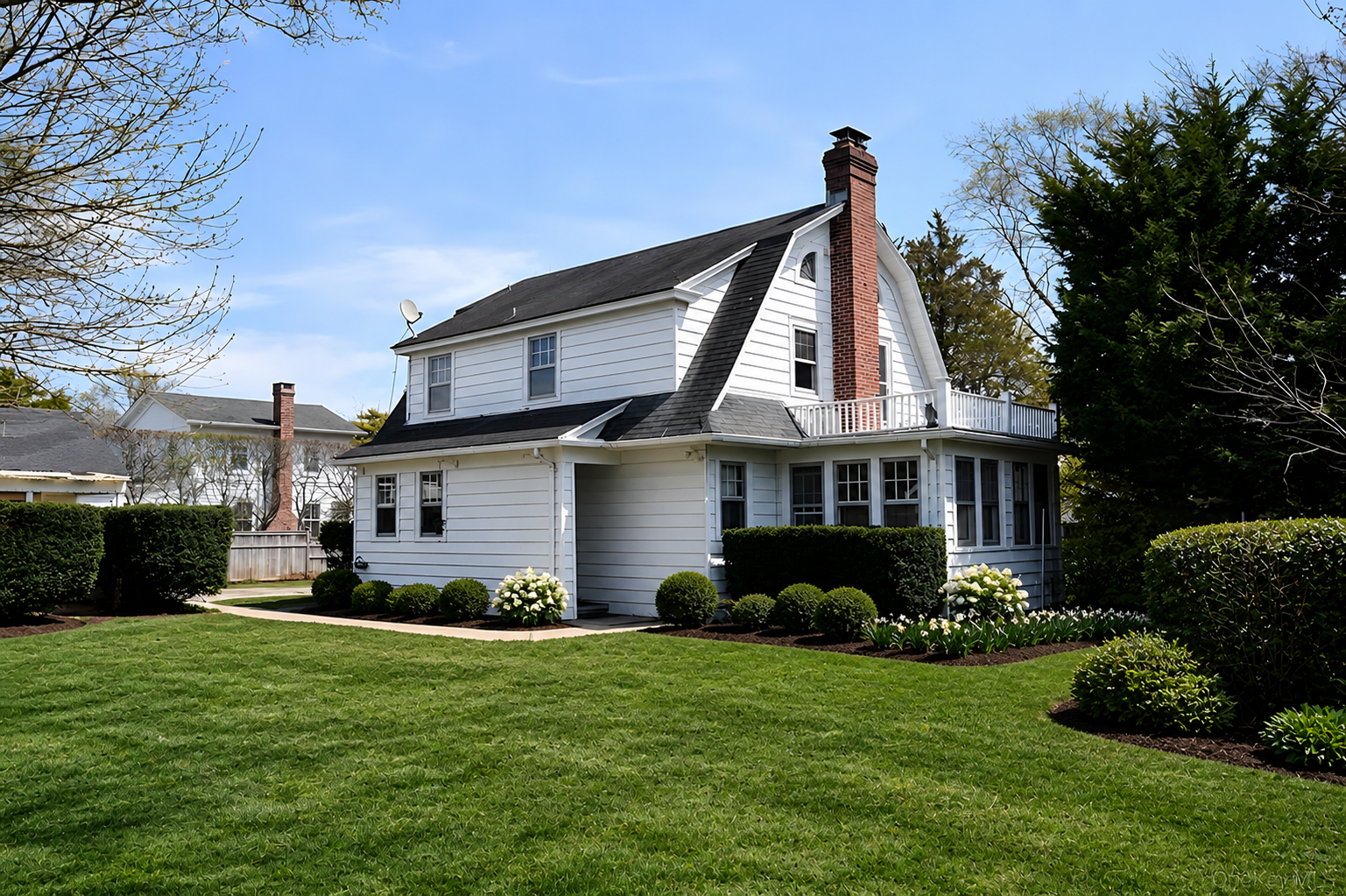 A white two-story house with a black roof and red brick chimney, surrounded by a well-maintained green lawn and bushes, on a clear day with blue sky.