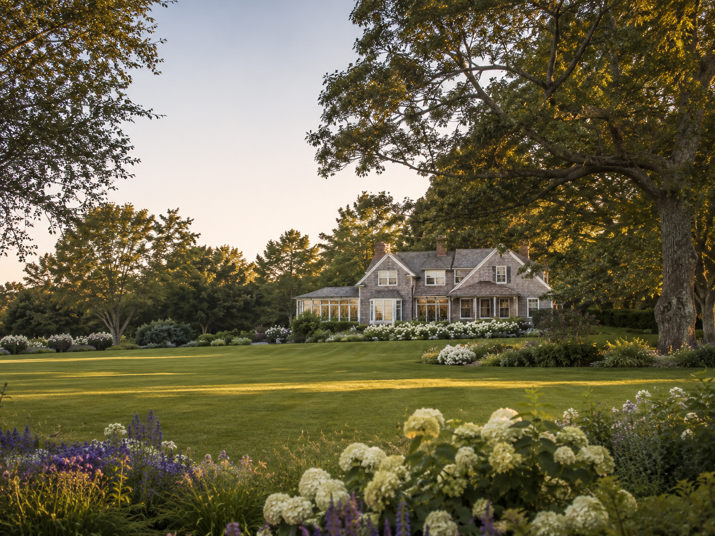 A large house with gray shingle siding and multiple gables, surrounded by well-maintained lawns and gardens, with tall trees and a clear sky in the background.