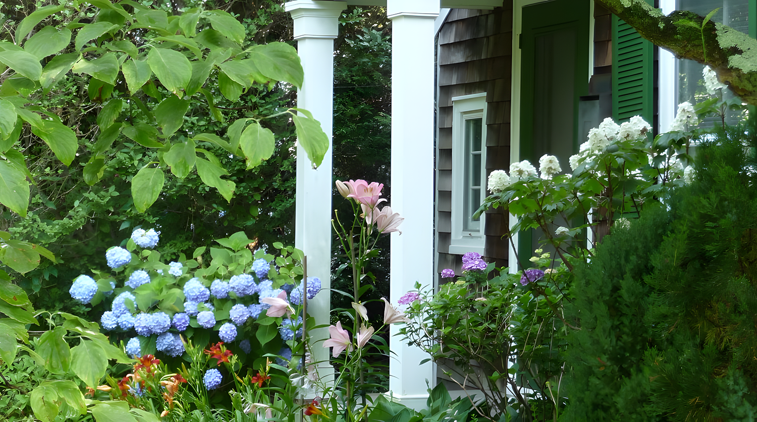 A garden scene with various colorful flowers, green foliage, a house with wooden siding, white columns, a window, and a green shutter.
