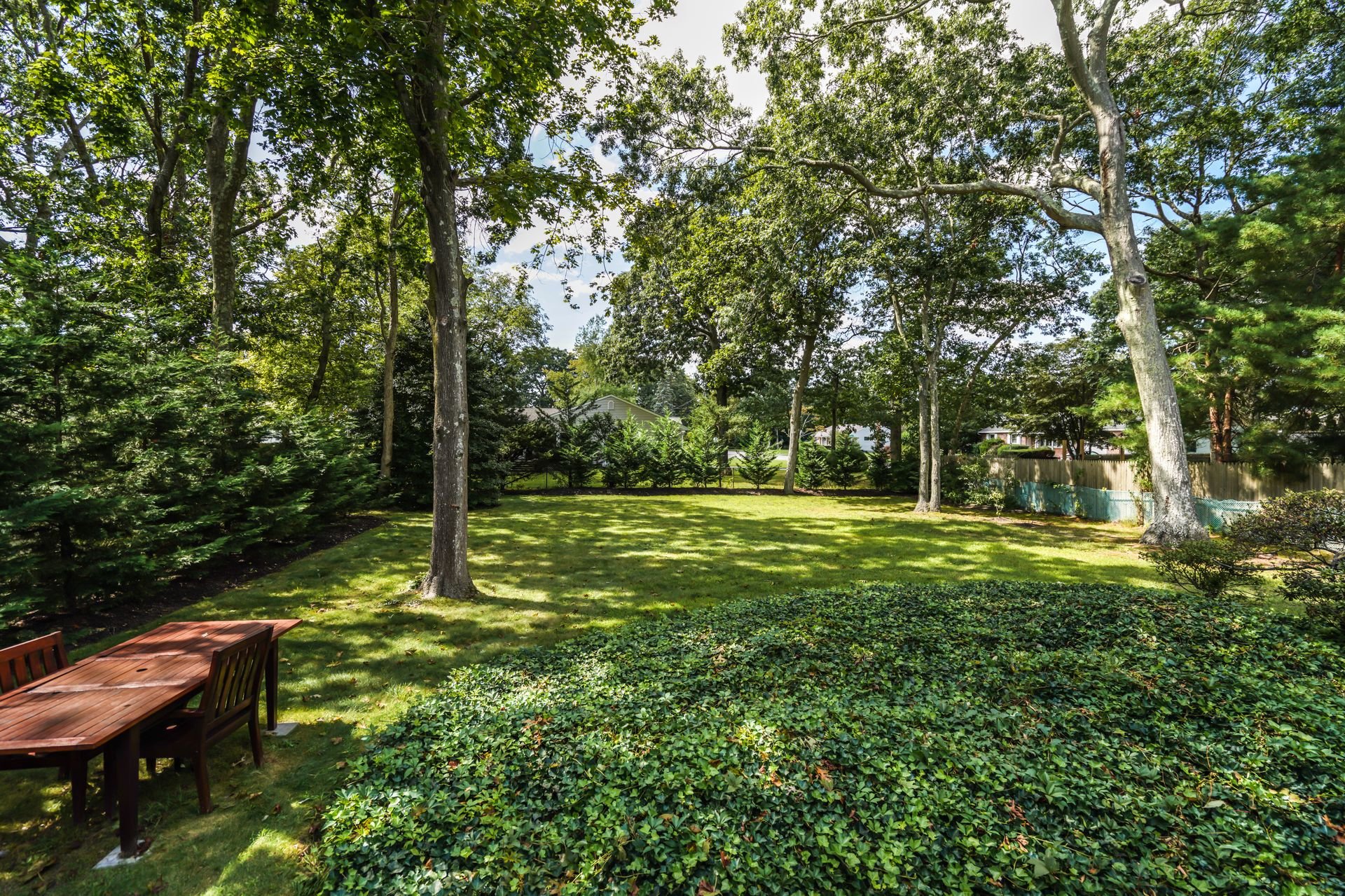 A lush backyard with tall trees, a well-manicured lawn, and surrounding shrubs. There's a wooden outdoor table with chairs in the foreground and a wooden fence with neighboring houses in the background.