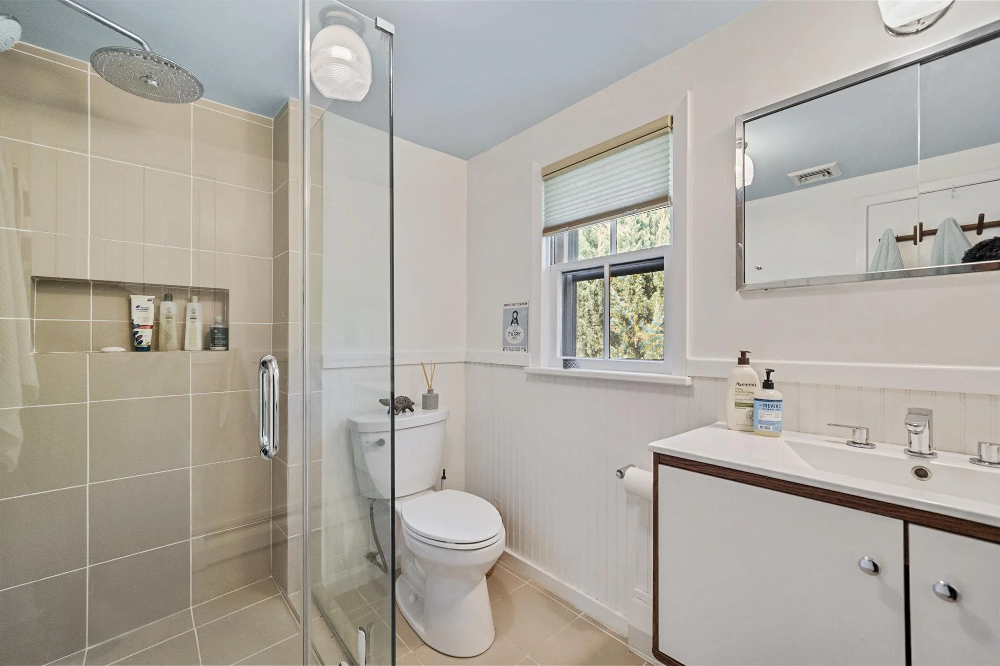 A clean bathroom with a walk-in shower with beige tiles, a glass door, and a recessed shelf with toiletries. There is a white toilet next to the shower, a window with blinds, and a mirror above the sink. The sink has a white countertop and drawers wi