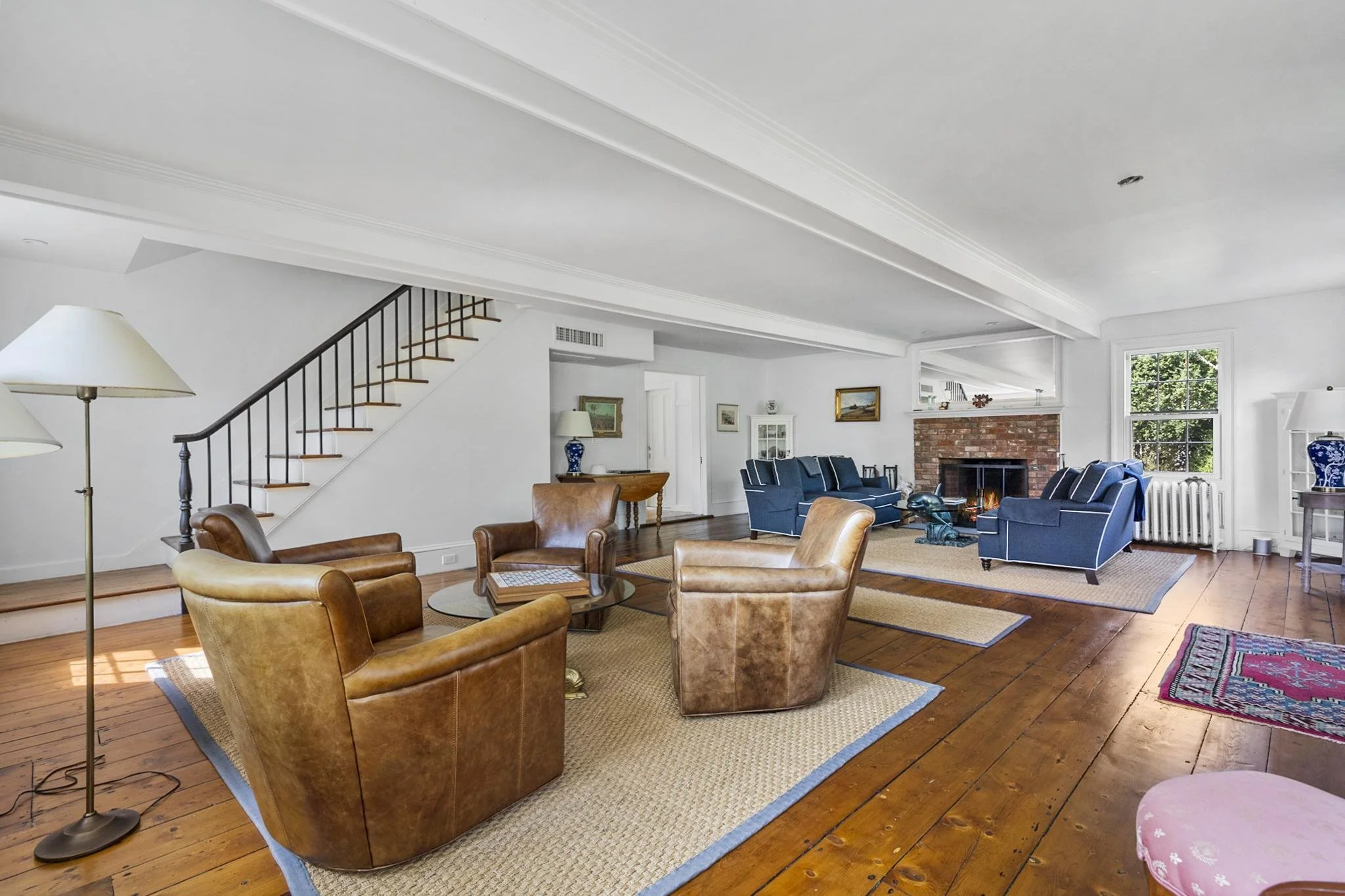 A spacious living room with wooden floors and white walls, furnished with brown leather armchairs and blue upholstered couches. There is a brick fireplace, large windows with greenery outside, and art in frames on the walls. A staircase with a black 