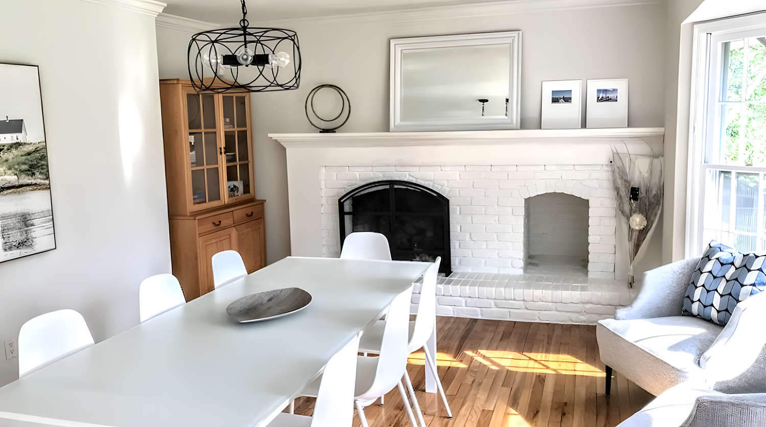 Living room with white brick fireplace, wooden dining table and white chairs, grey armchair with patterned pillow, large window with view of outside greenery, and decorative elements on the mantel.