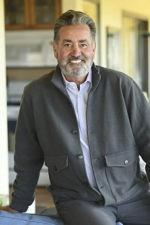 A smiling middle-aged man with gray hair and a beard, wearing a gray jacket and white shirt, standing indoors with a kitchen in the background.