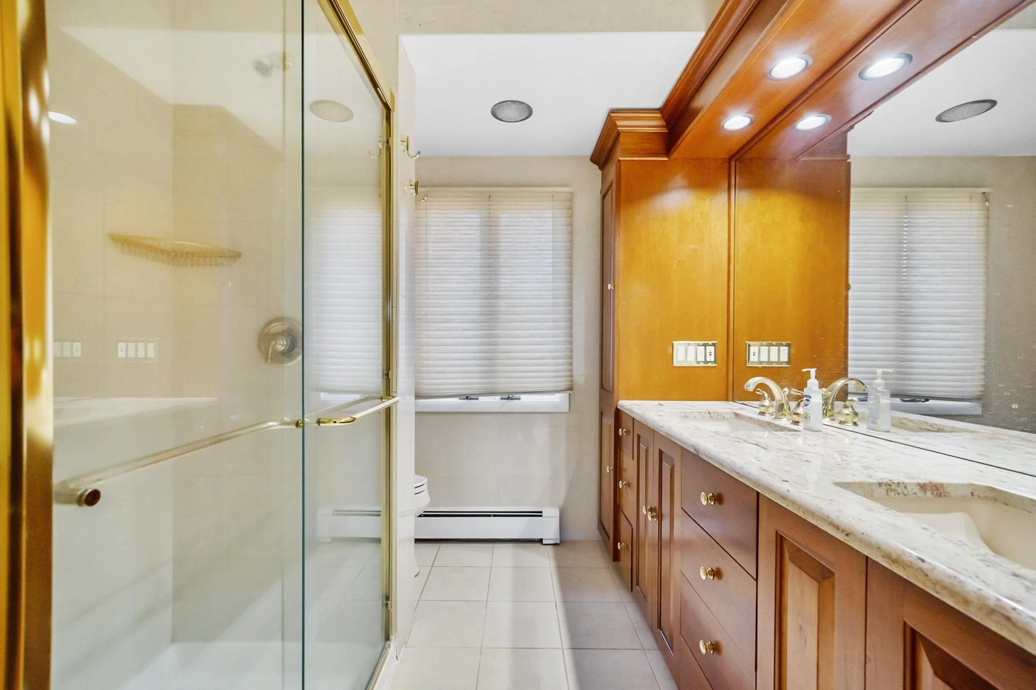 Bathroom with a double vanity sink, wooden cabinets, marble countertop, window with blinds, shower area with glass door, and various fixtures