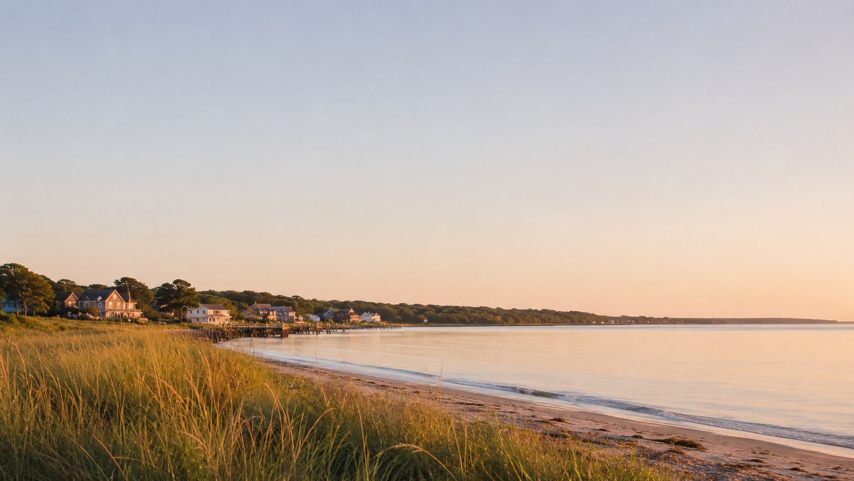 A serene beach scene during sunset with sandy shoreline, calm water, and a row of houses along the coast in the distance, some on piers extending into the water, with grassy dunes in the foreground.
