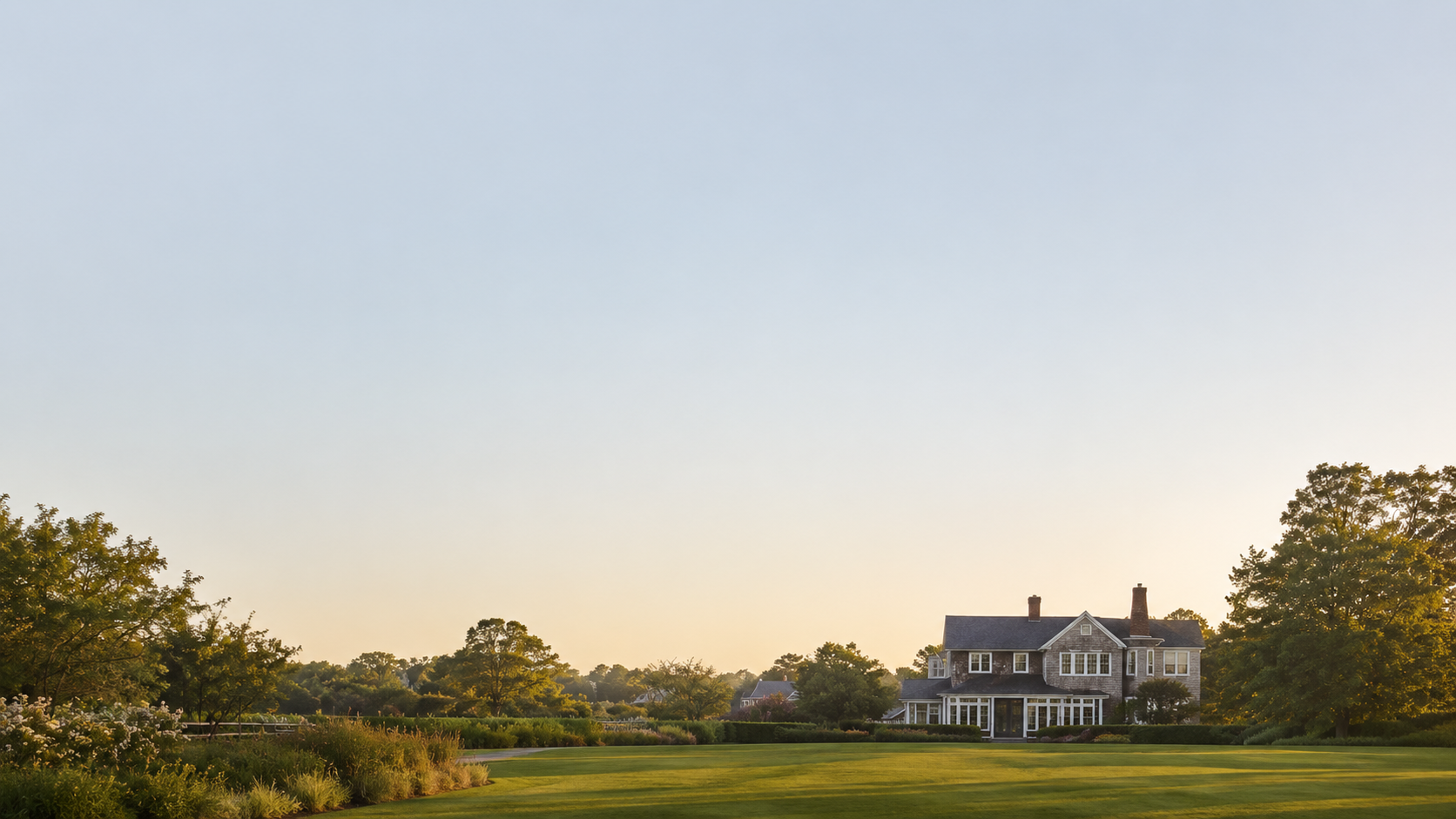 A large historic house with a wraparound porch and multiple chimneys, surrounded by well-maintained lawn and trees at sunset.