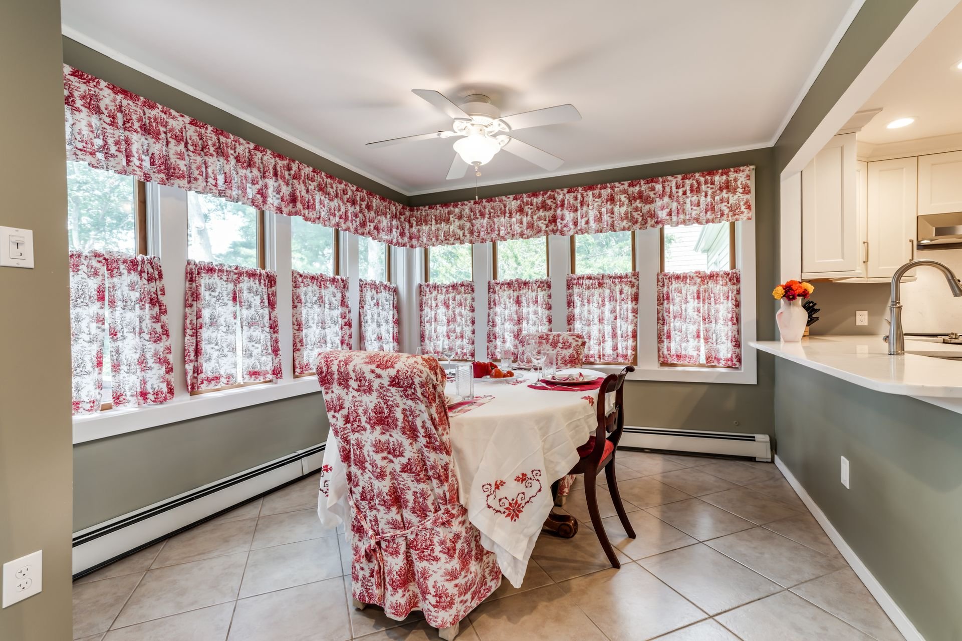 A dining area with a table covered by a white tablecloth and surrounded by chairs with red and white toile fabric covers. The windows have matching red and white toile curtains and valances. The room has a ceiling fan with a light, green painted wall