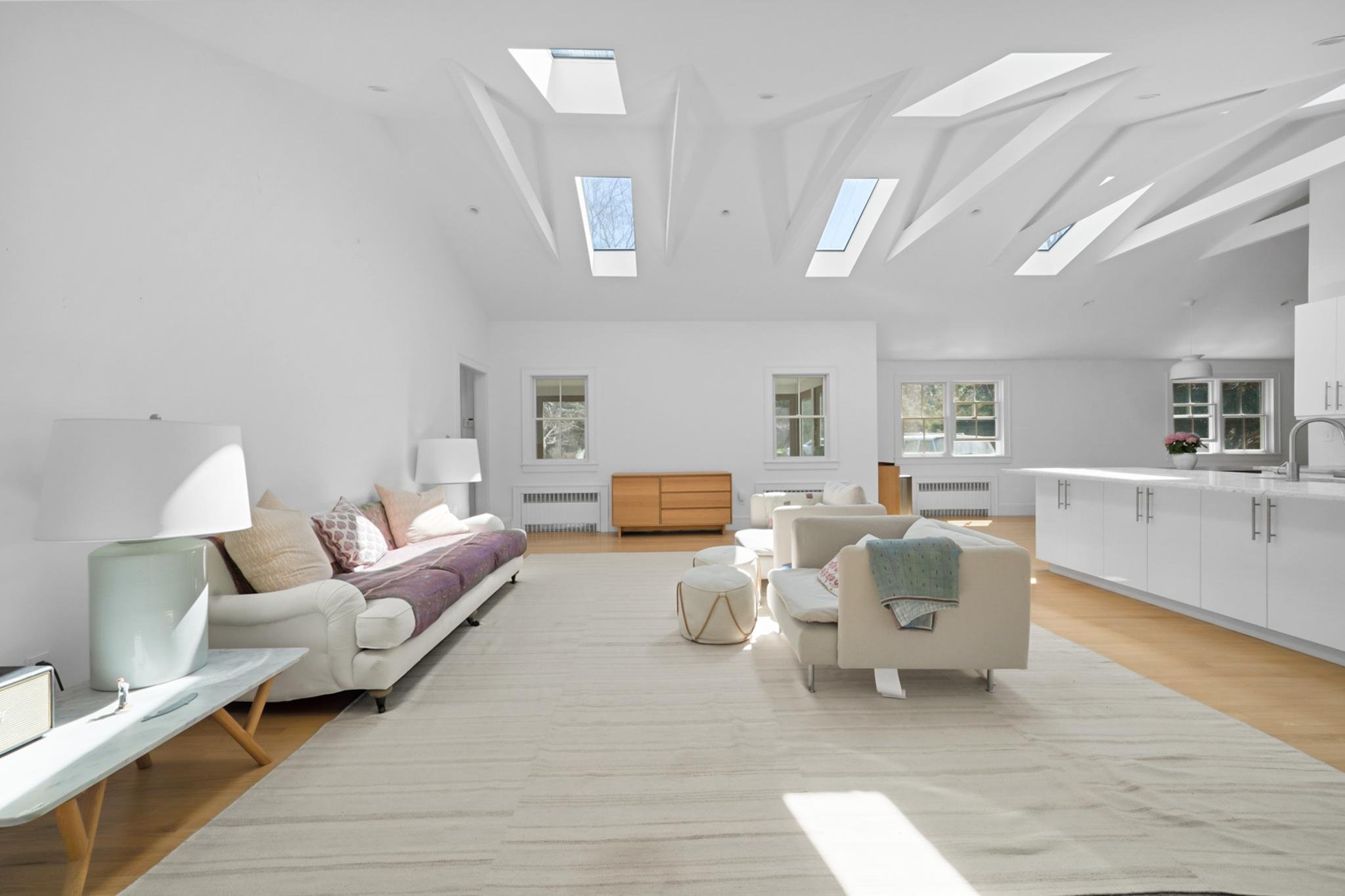 Bright, modern living room with white walls and ceiling, multiple rectangular skylights, a white sofa with beige and patterned pillows, a coffee table, a white chair, a wooden console, and a kitchen area with white cabinets and a pink potted plant.