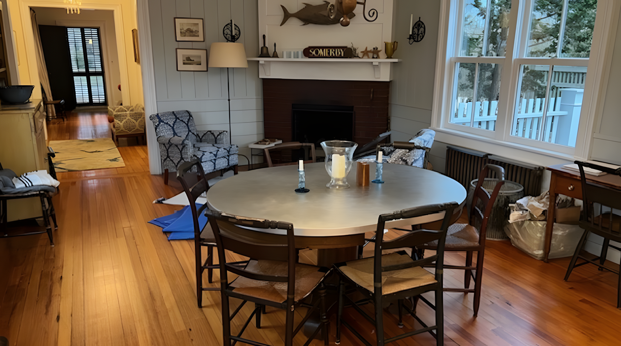 Living room with a round dining table surrounded by chairs, a fireplace with a mantle decorated with various items, large windows on the right, and a hallway leading to a back door on the left. The room has hardwood floors and various decorative elem