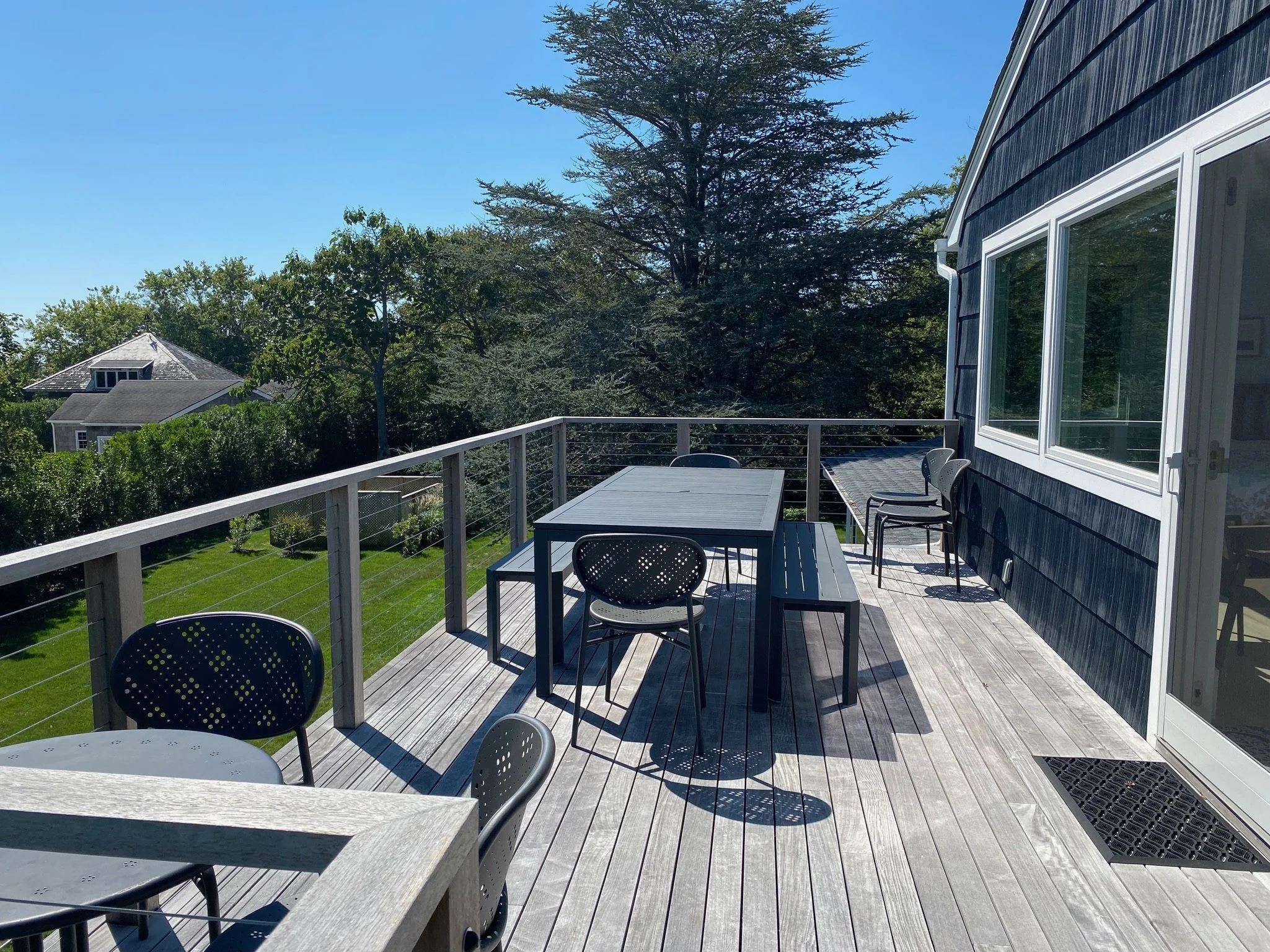 An outdoor wooden deck with tables and chairs, overlooking a yard with lush green grass and trees under a clear blue sky.