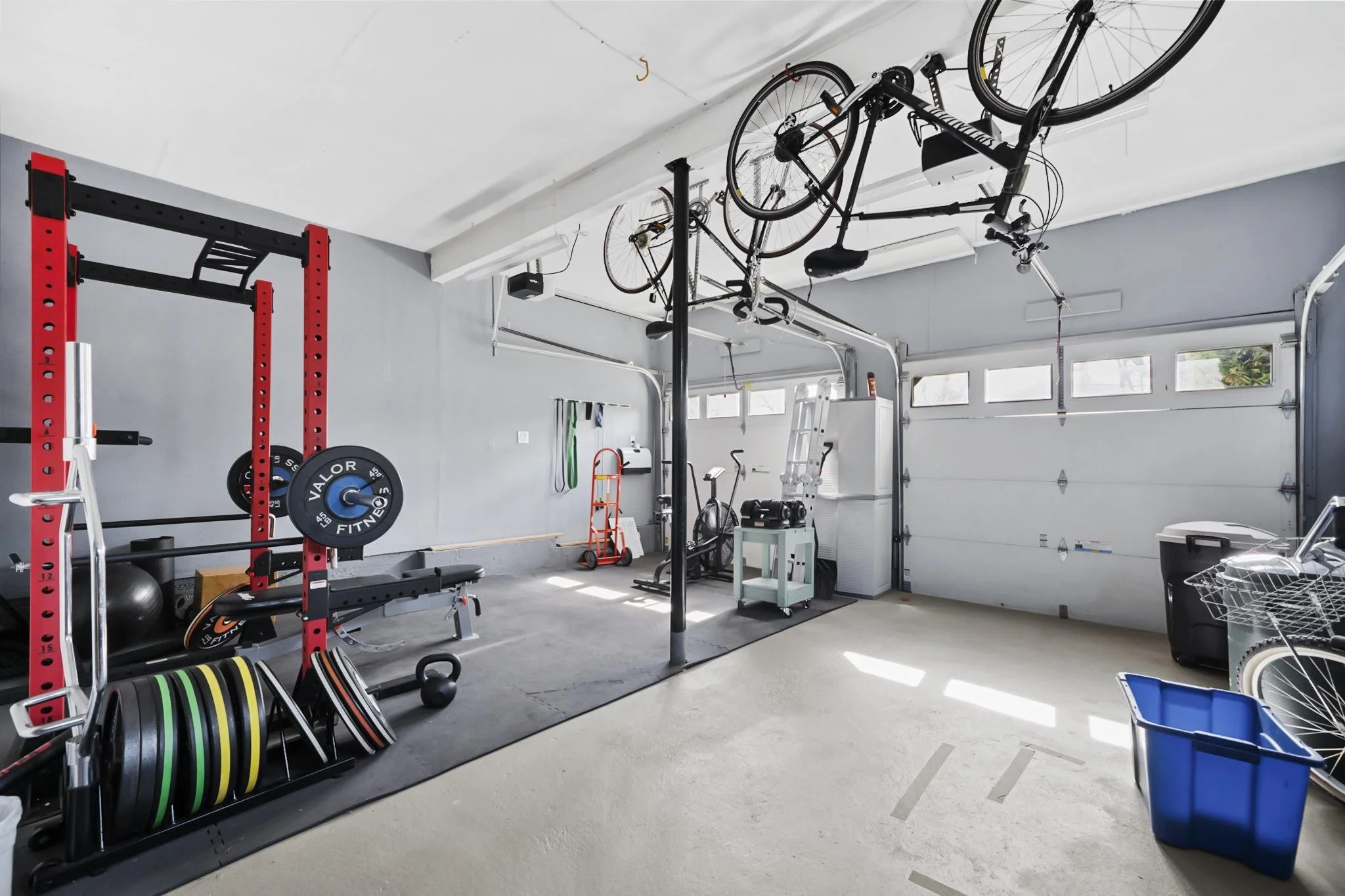 Home gym in a garage with exercise equipment, a squat rack, weight plates, a boxing bag, bicycles mounted on the ceiling, and various fitness accessories.