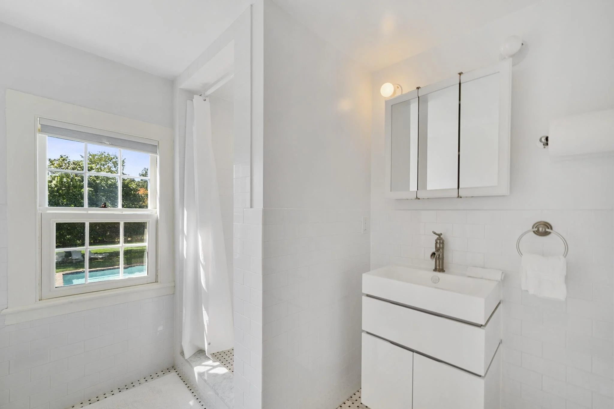 A bright, white bathroom with a large window showing trees outside. The bathroom has white tile walls, a white vanity with a faucet, a mirror, and ceiling lighting. There is a shower area with a white curtain and white tile floor.