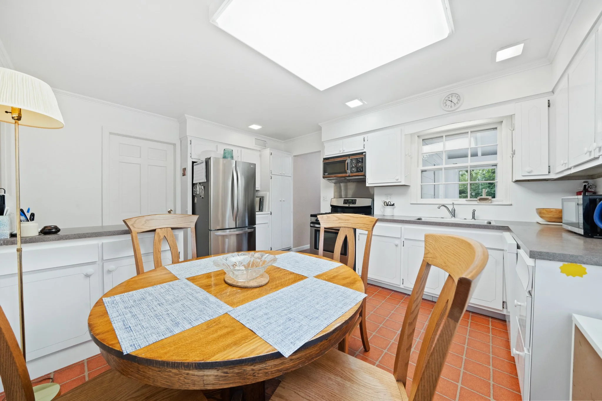 A kitchen with white cabinets, stainless steel refrigerator, microwave, and oven; a round wooden dining table with a blue and white table runner, surrounded by wooden chairs; a window with a view outside; and various kitchen appliances and decor.