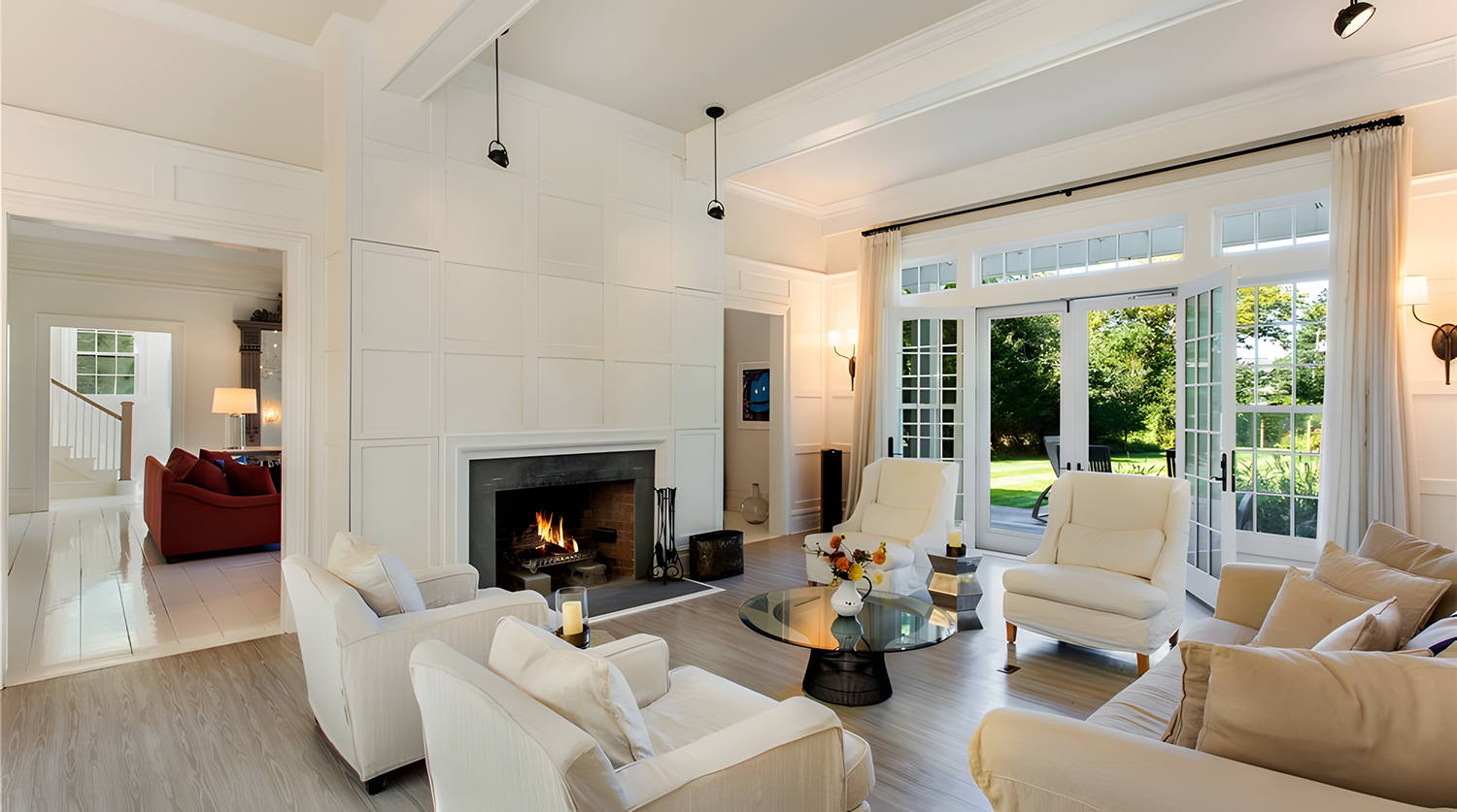 Bright living room with white walls and furniture, fireplace, glass doors opening to a green outdoor lawn, and sunlight streaming in.
