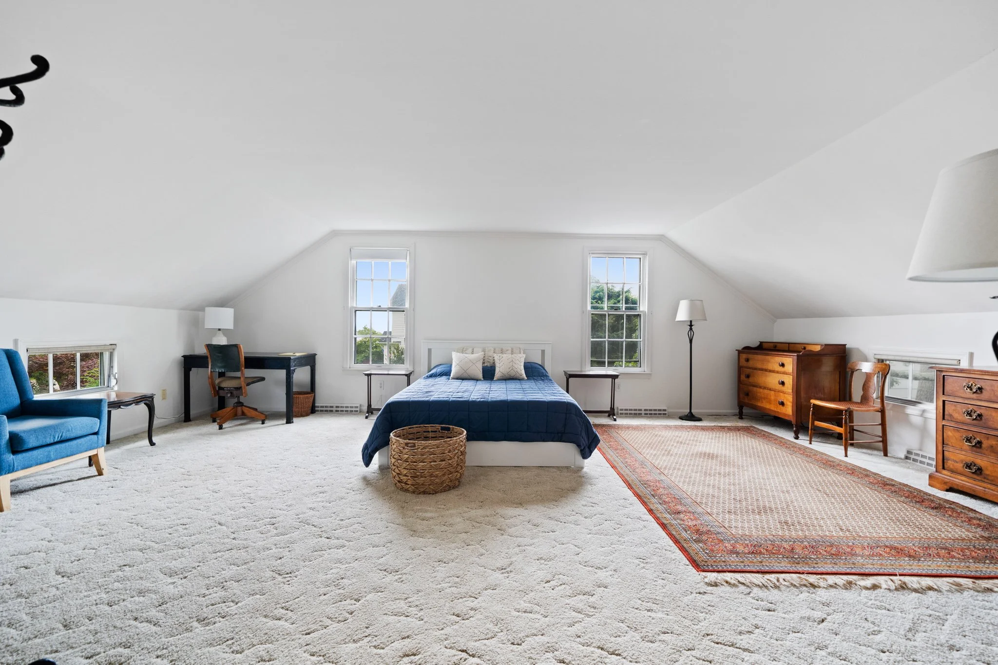 A spacious bedroom with two windows, a bed with a navy blue cover and white pillows, blue armchair, black writing desk with chair, side tables, a wooden dresser, a standing lamp, and a woven basket, with a beige carpet and a patterned rug.