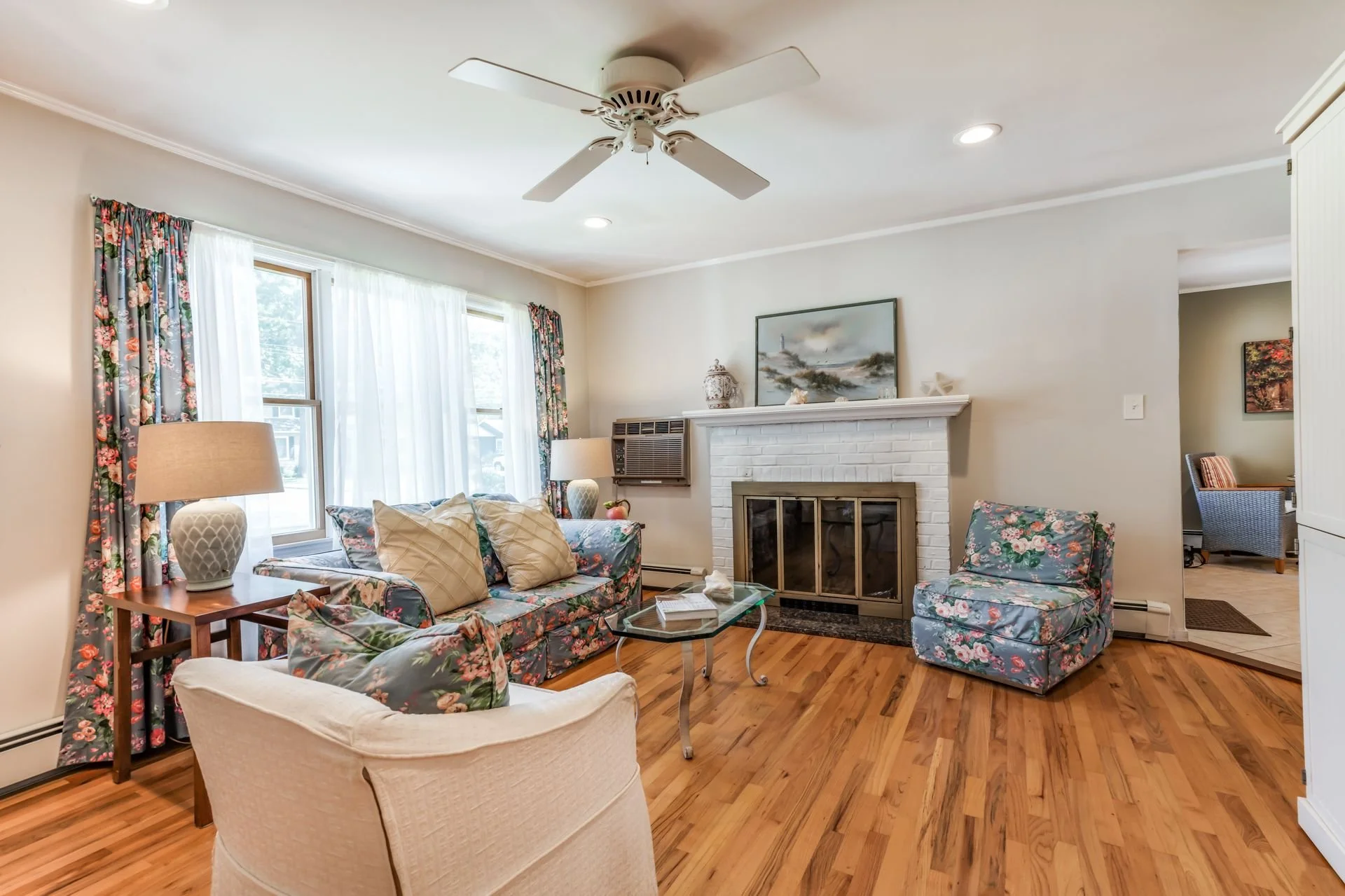 Living room with floral patterned couch and armchair, a white brick fireplace, a glass coffee table, window with floral curtains, a side table with a lamp, hardwood flooring, and a ceiling fan.