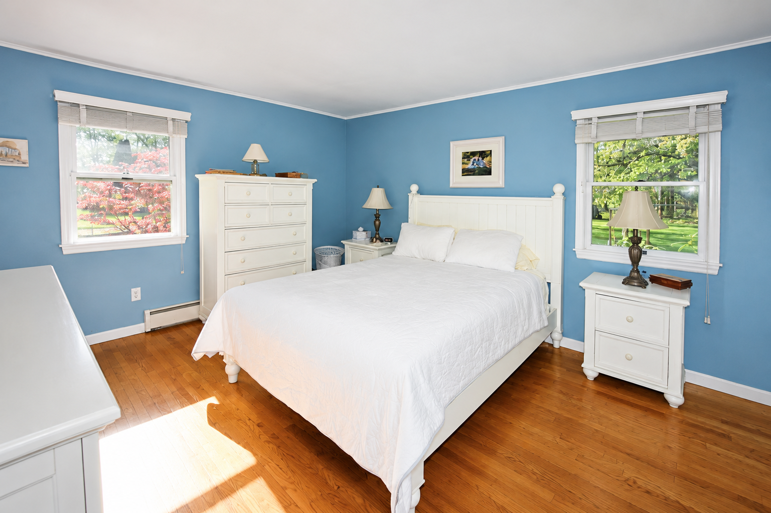 A bedroom with blue walls, white furniture, a white bed with pillows, two windows with white blinds showing trees outside, two nightstands with lamps, a tall white dresser, and a small framed picture above the bed.