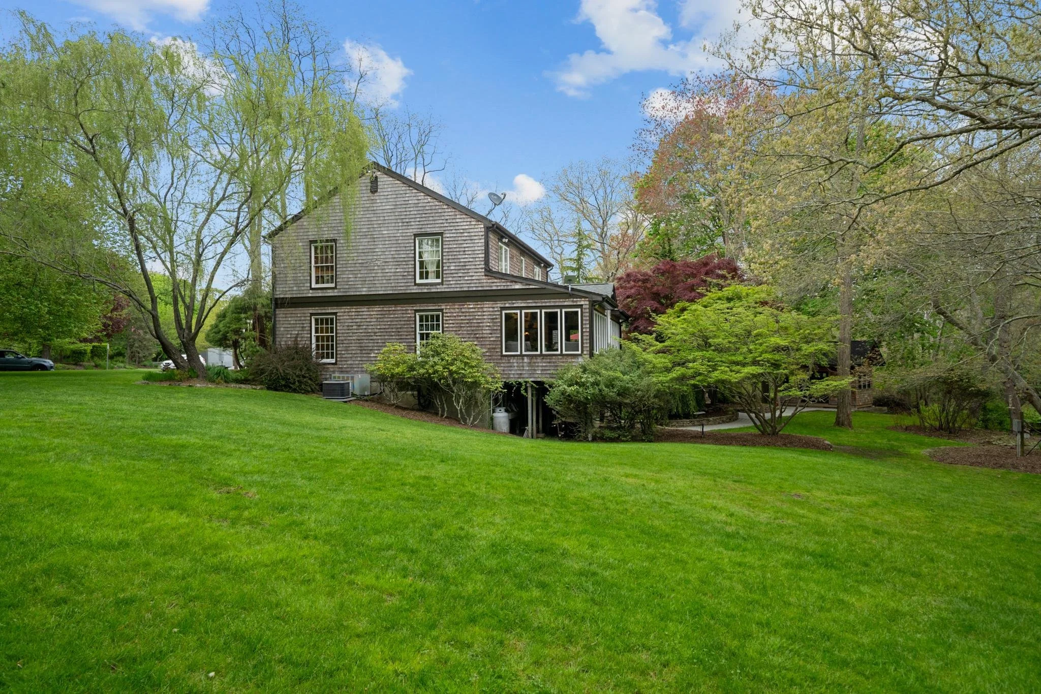 A two-story house with wooden shingles in a lush green yard and tall trees, some with budding leaves, under a bright blue sky with white clouds.