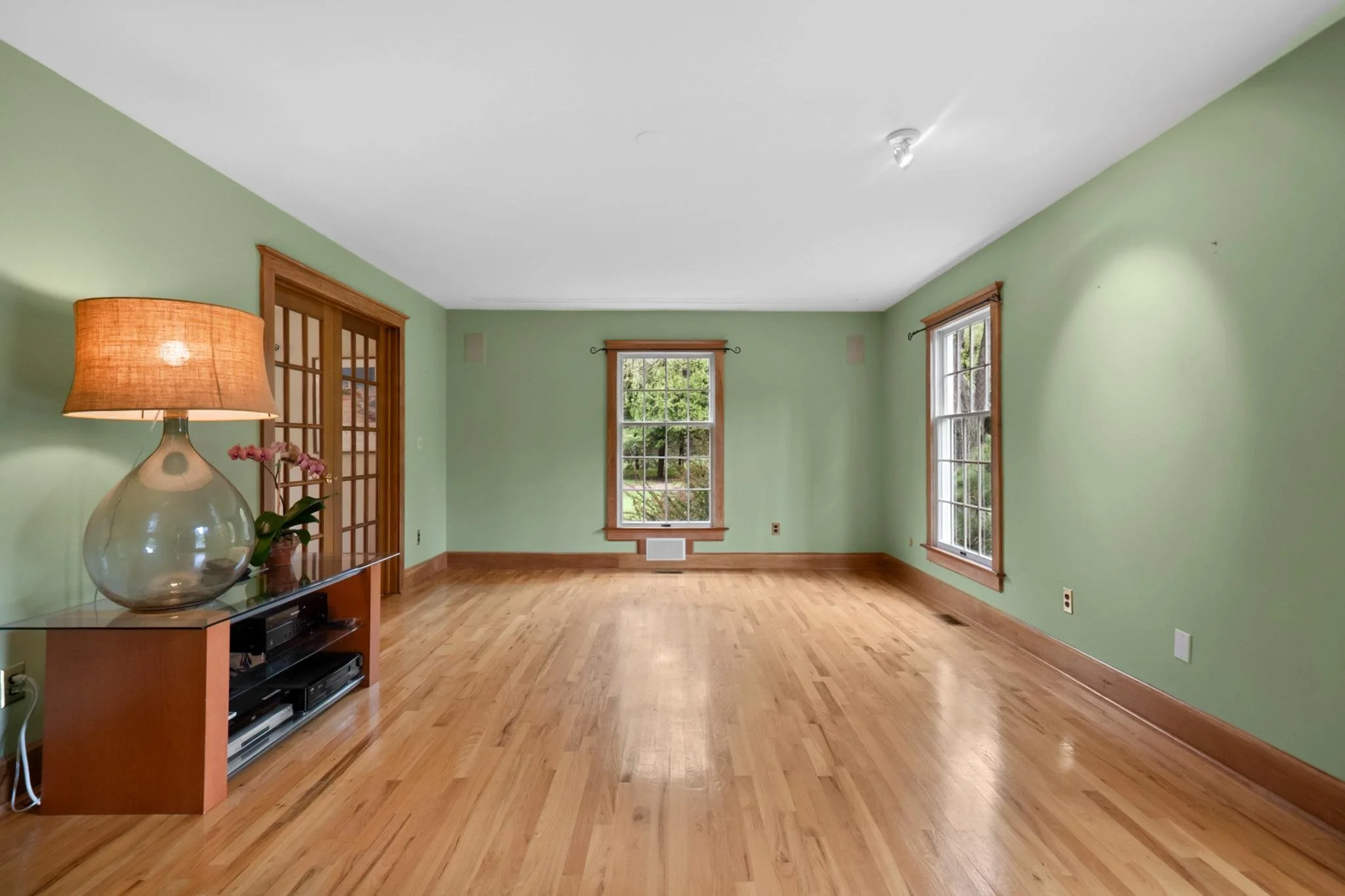 Empty living room with green walls, hardwood floors, and large windows letting in natural light; a lamp and entertainment center are visible.