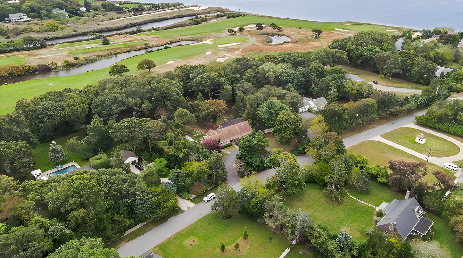 Aerial view of a suburban neighborhood with lush green trees, houses, swimming pool, winding roads, and a golf course with water features in the background.