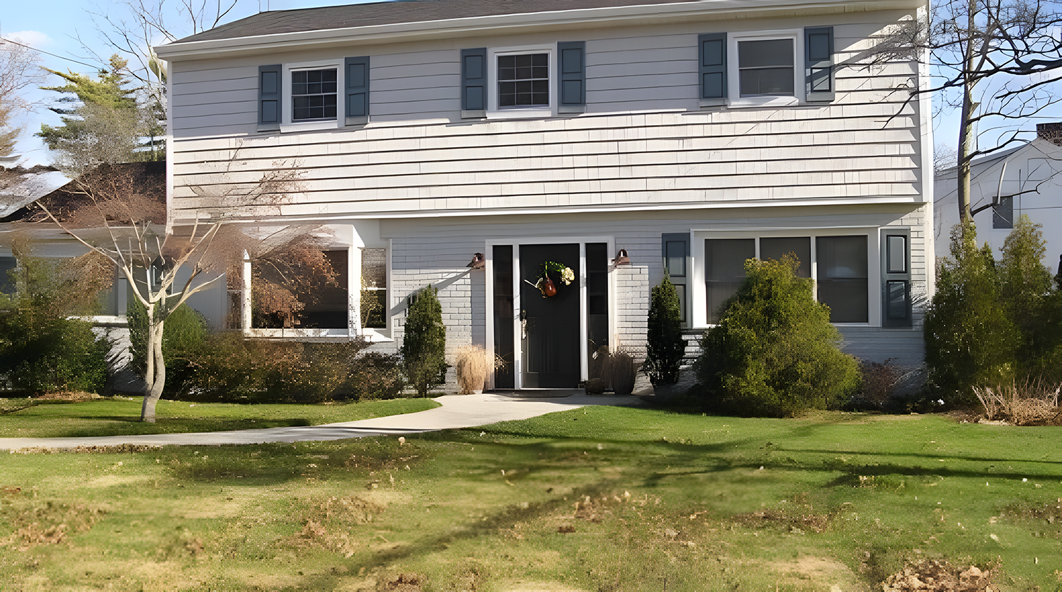Front view of a two-story house with white siding, dark blue shutters, large windows, and a black front door decorated with a wreath and a watering can. The house is surrounded by a well-maintained lawn, bushes, and trees, with a concrete pathway lea