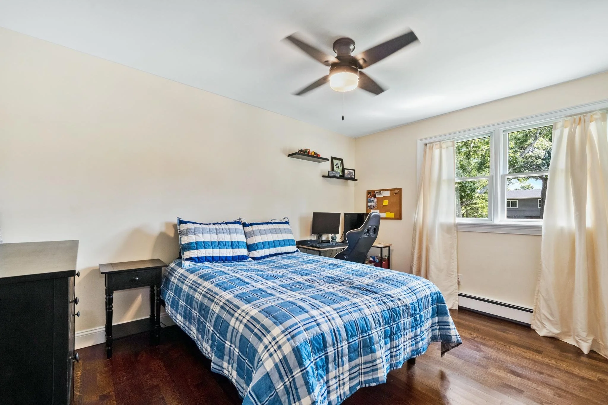 A bedroom with a bed featuring blue and white plaid bedding, two black nightstands, a desk with a computer and gaming chair, a wall shelf with decor, a corkboard, a window with beige curtains, a ceiling fan, and hardwood floors.