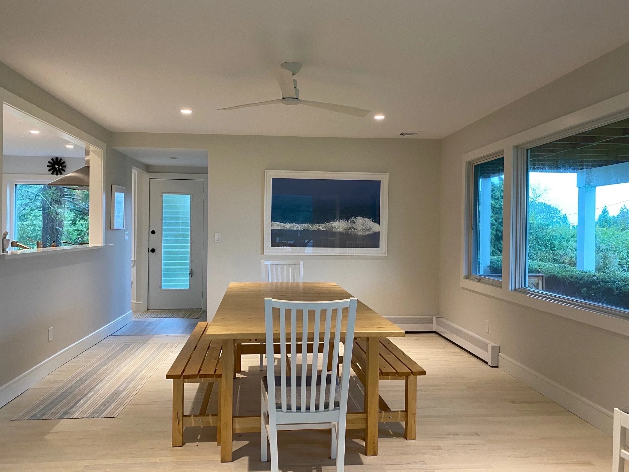 A dining room with wooden table and chairs, large windows showing trees outside, a framed artwork on the wall, and a ceiling fan.