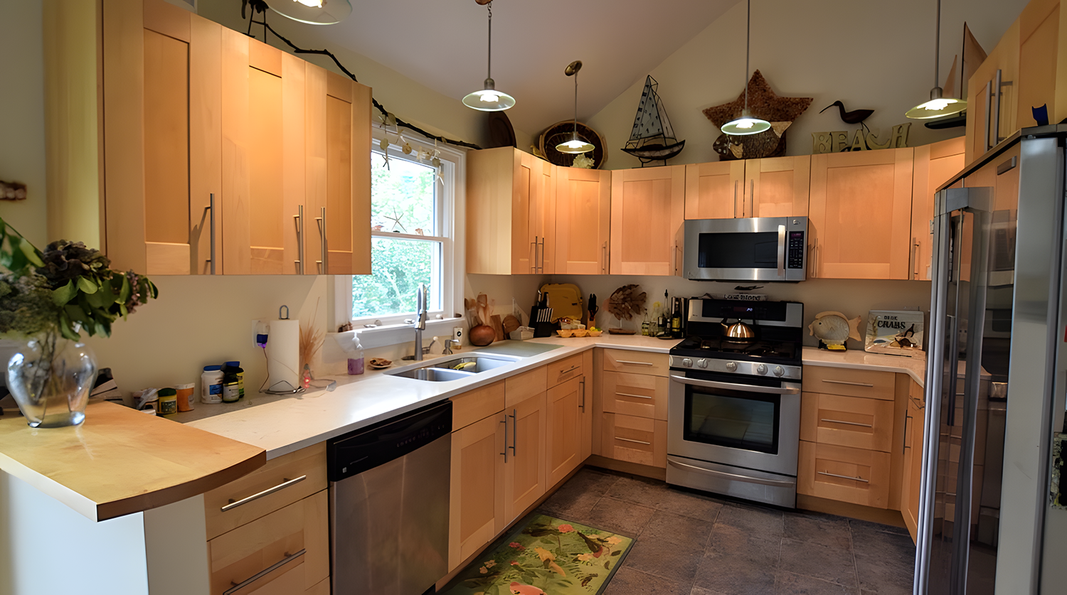 A kitchen with light wooden cabinets, white countertops, a window above the sink, stainless steel appliances including a microwave, oven, dishwasher, and refrigerator, various decorative items on top of the cabinets, and a green floral rug on the til