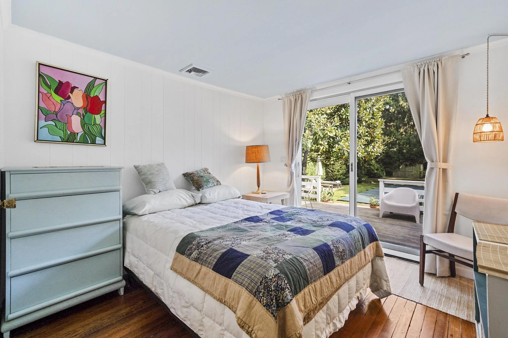 A cozy bedroom with a bed covered in a patchwork quilt, a light blue chest of drawers, a framed floral artwork on the wall, a floor lamp, and sliding glass doors leading to a backyard with trees and outdoor furniture.