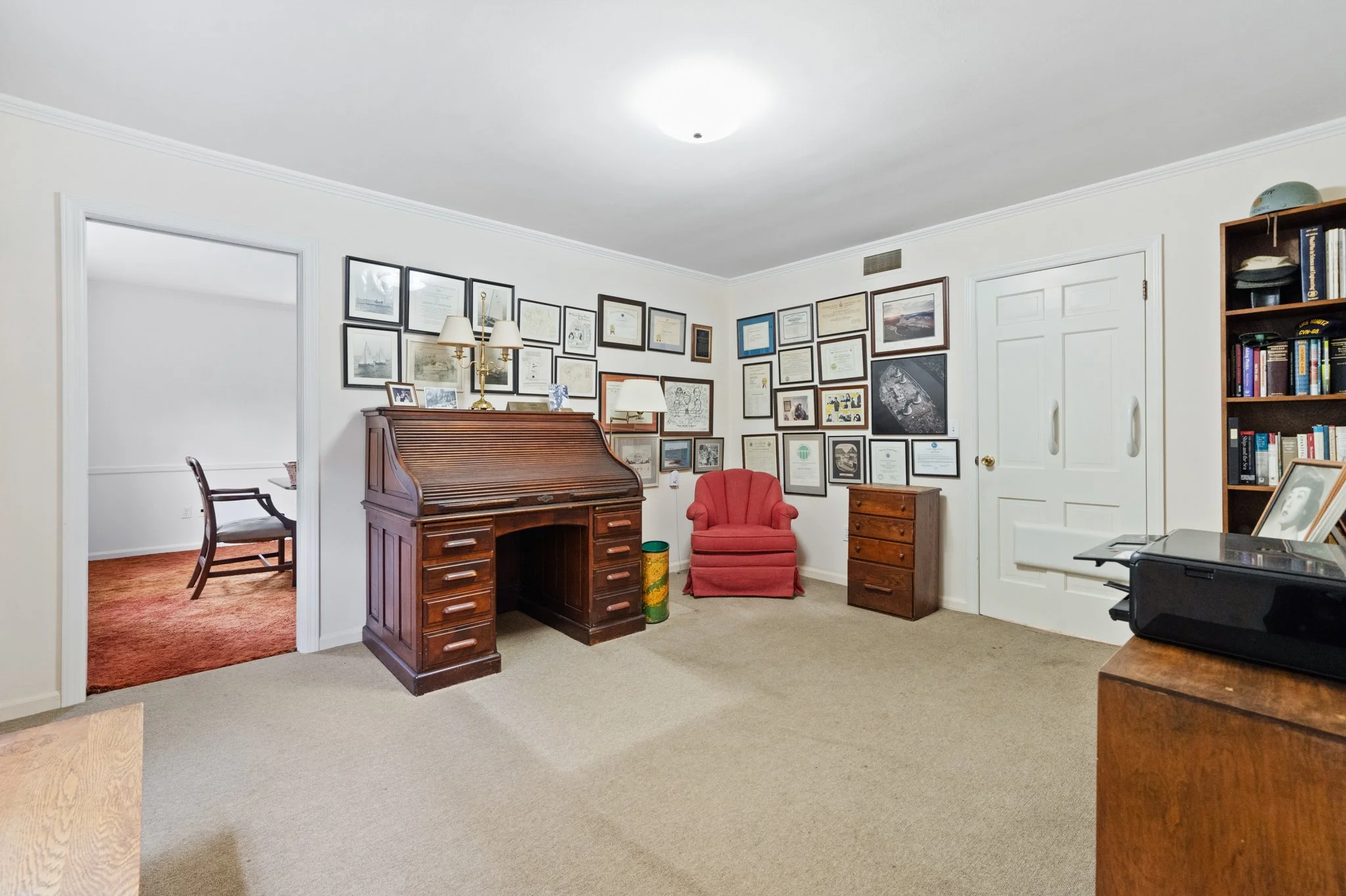 Living room with a wall of framed certificates, a wooden secretary desk, a red armchair, and bookshelves, with a doorway leading to a carpeted room.