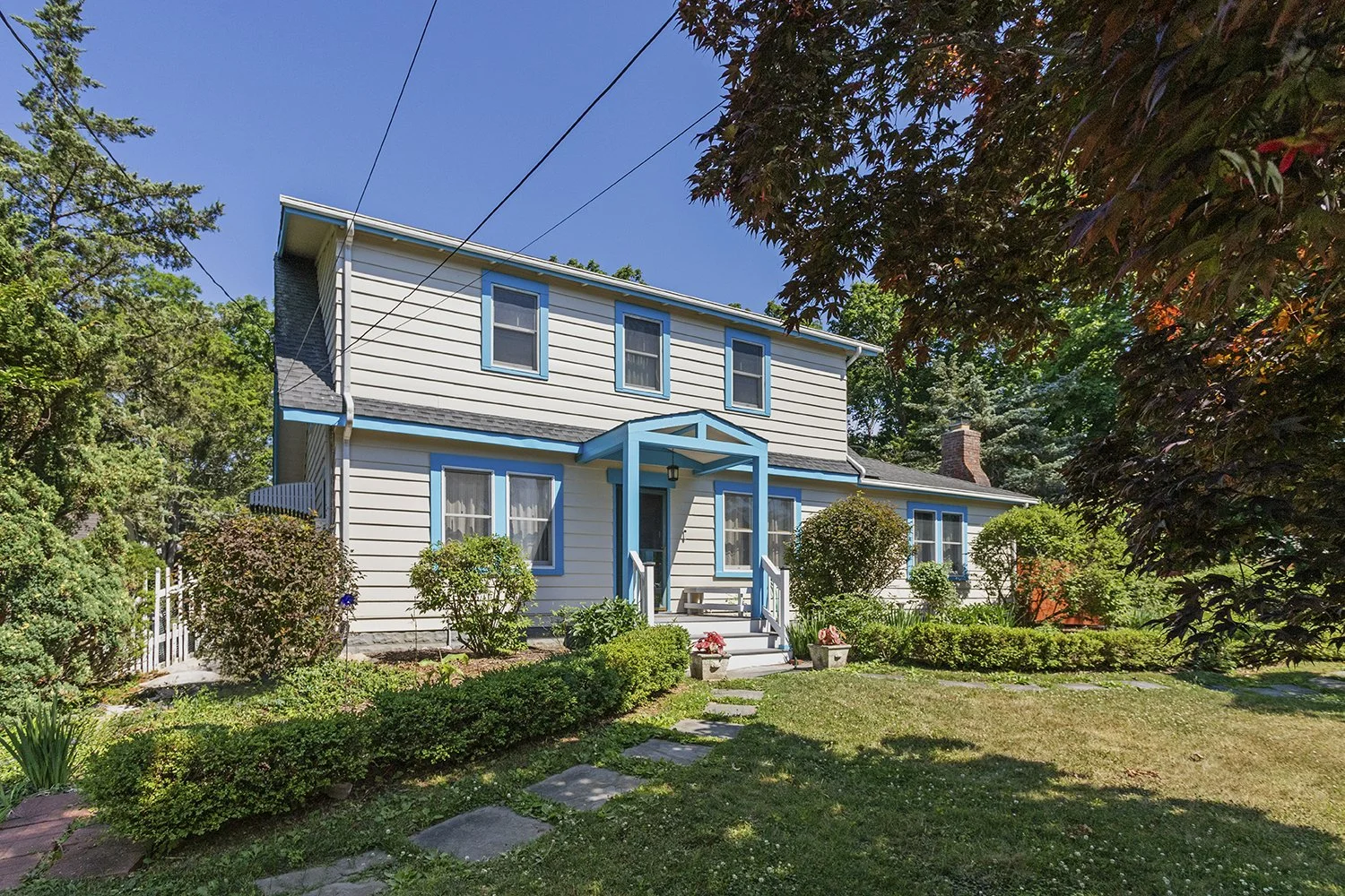 A two-story house with white siding, blue trim, and a small front porch with steps, surrounded by a well-maintained garden with bushes, flowering plants, and grass, under a clear blue sky.