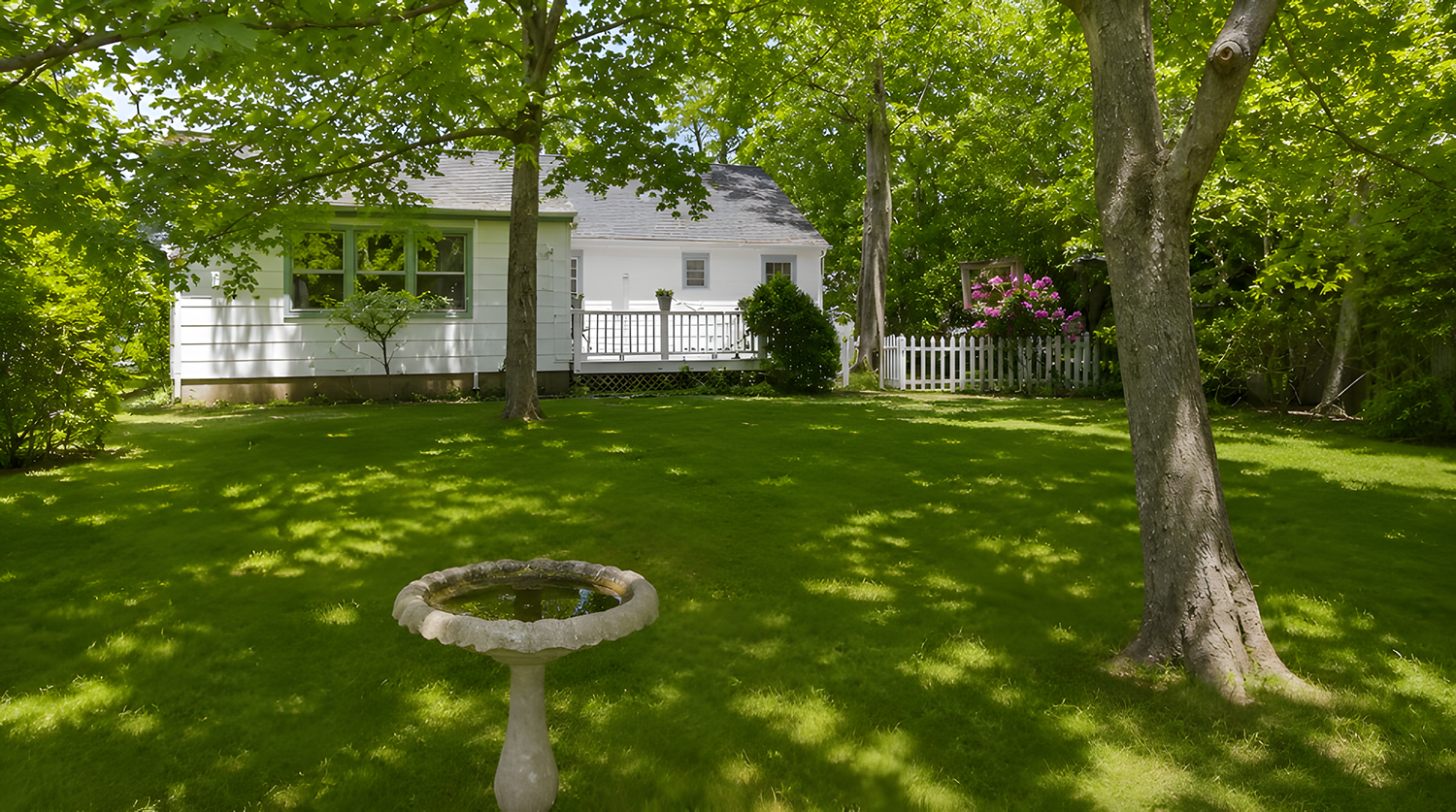 A backyard with green grass, trees providing shade, a birdbath, and a white house with a small porch and a white picket fence in the background.