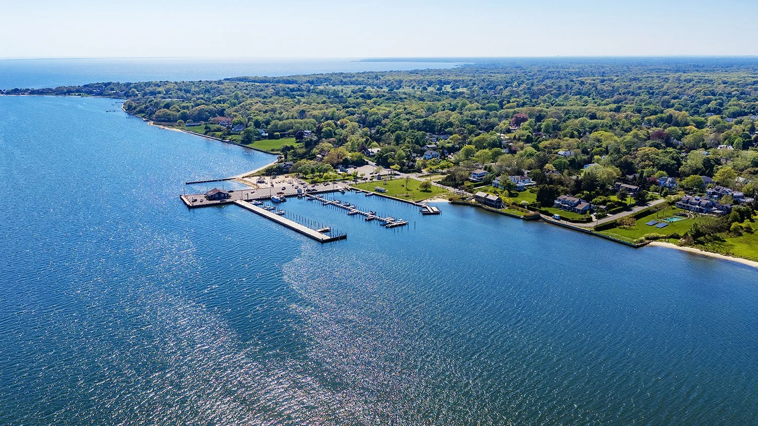 Aerial view of a coastal residential area with a marina, houses, trees, and a beach, next to a large body of water on a sunny day.