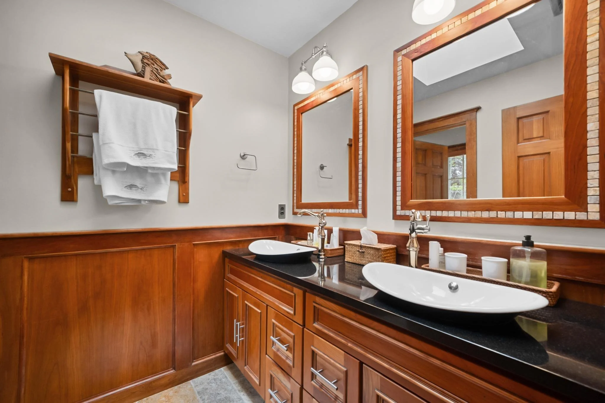 Bathroom with double mirrors, wooden cabinets, black countertop, vessel sinks, wall sconce lighting, towel rack, and toiletries.